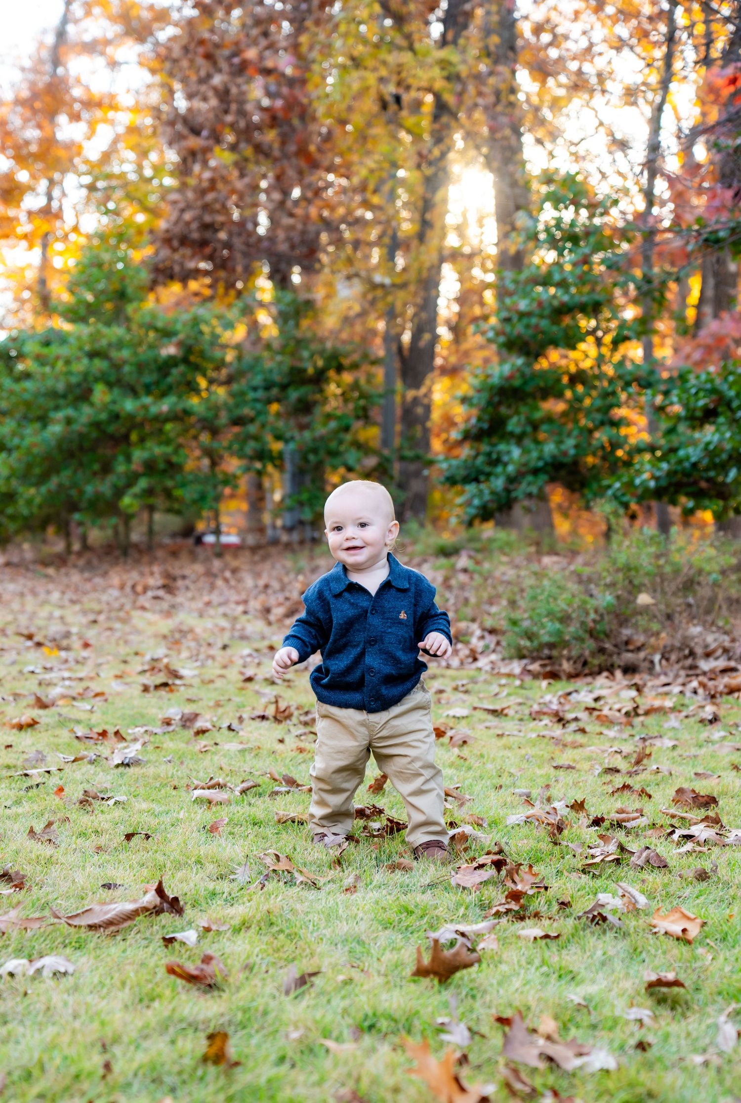 Baby in navy shirt and khaki pants walking on grass covered in autumn leaves during golden hour.