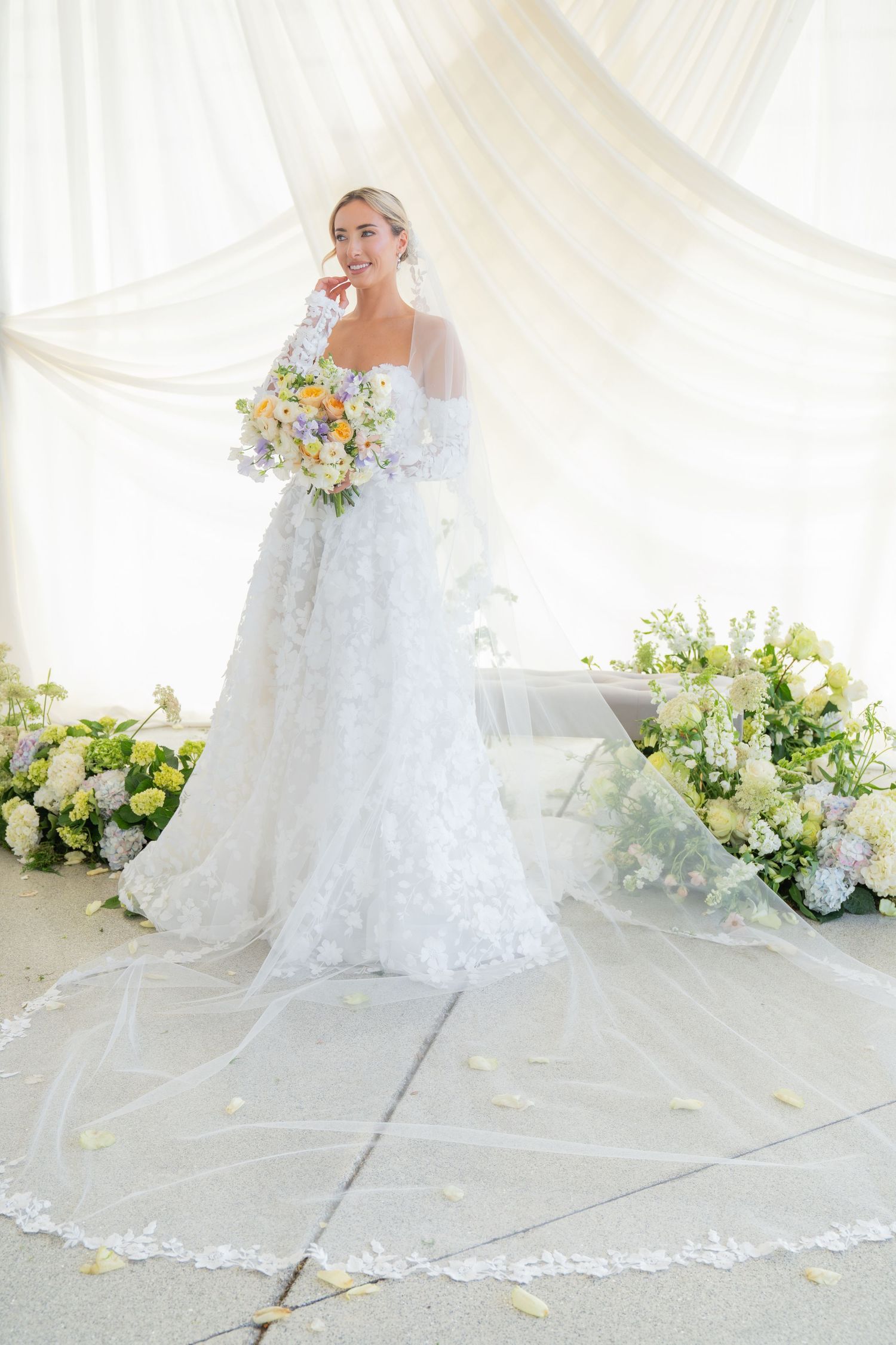 Bride wearing off-shoulder lace gown holds spring bouquet in white tent setting.