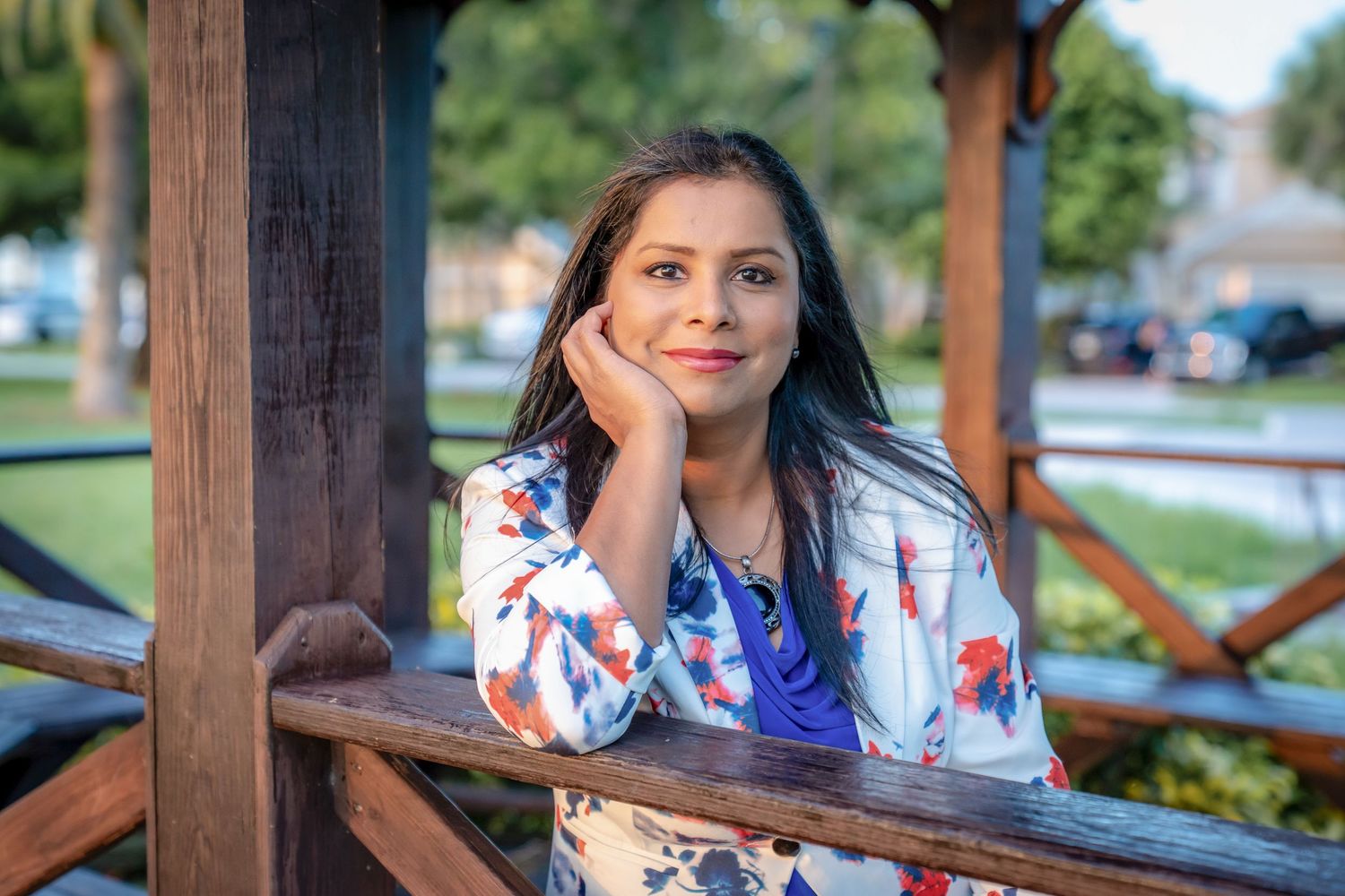 A person in a white floral dress poses in front of a wooden gazebo on a sunny day.