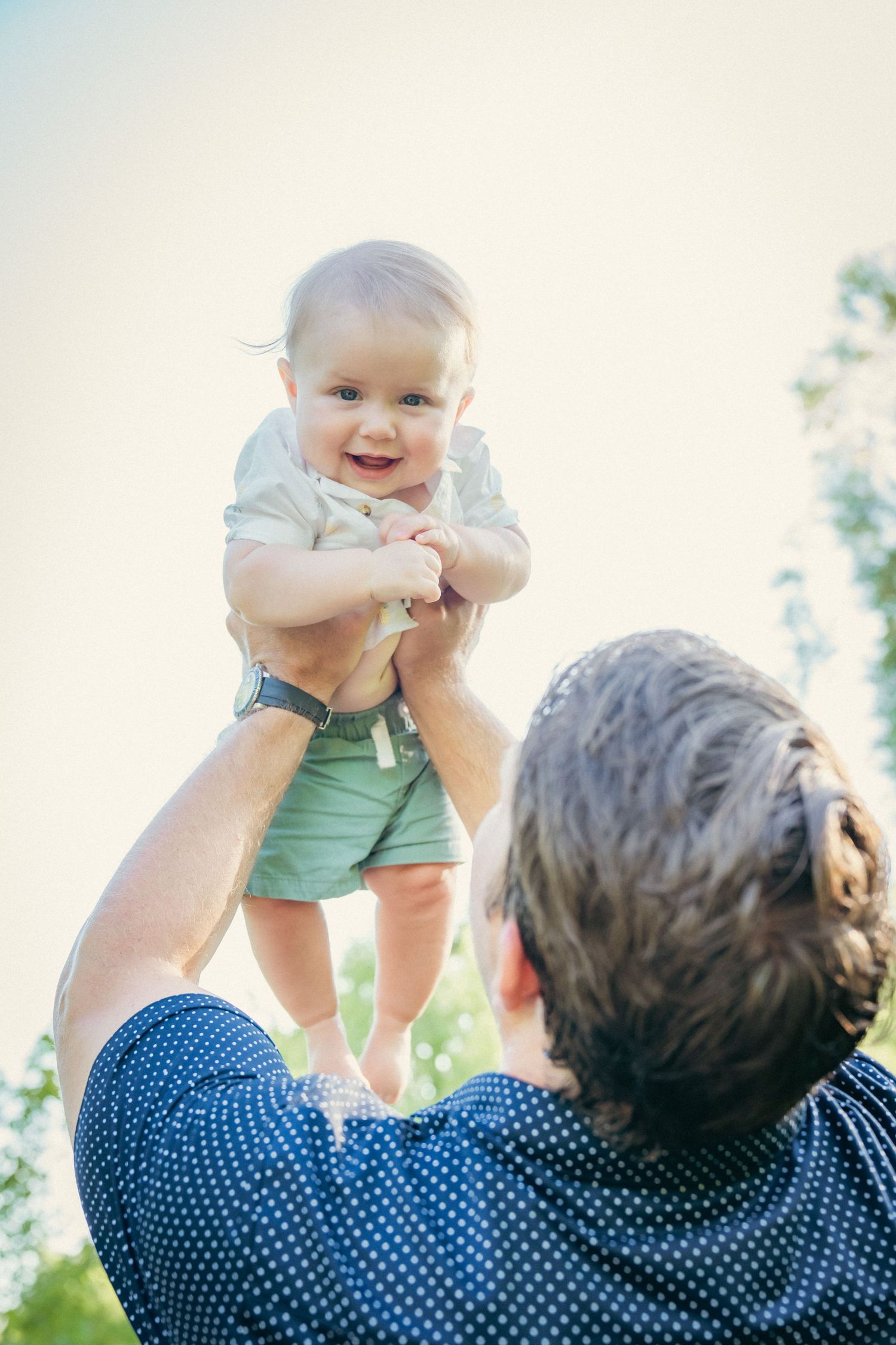 Baby being lifted playfully against bright sky background.