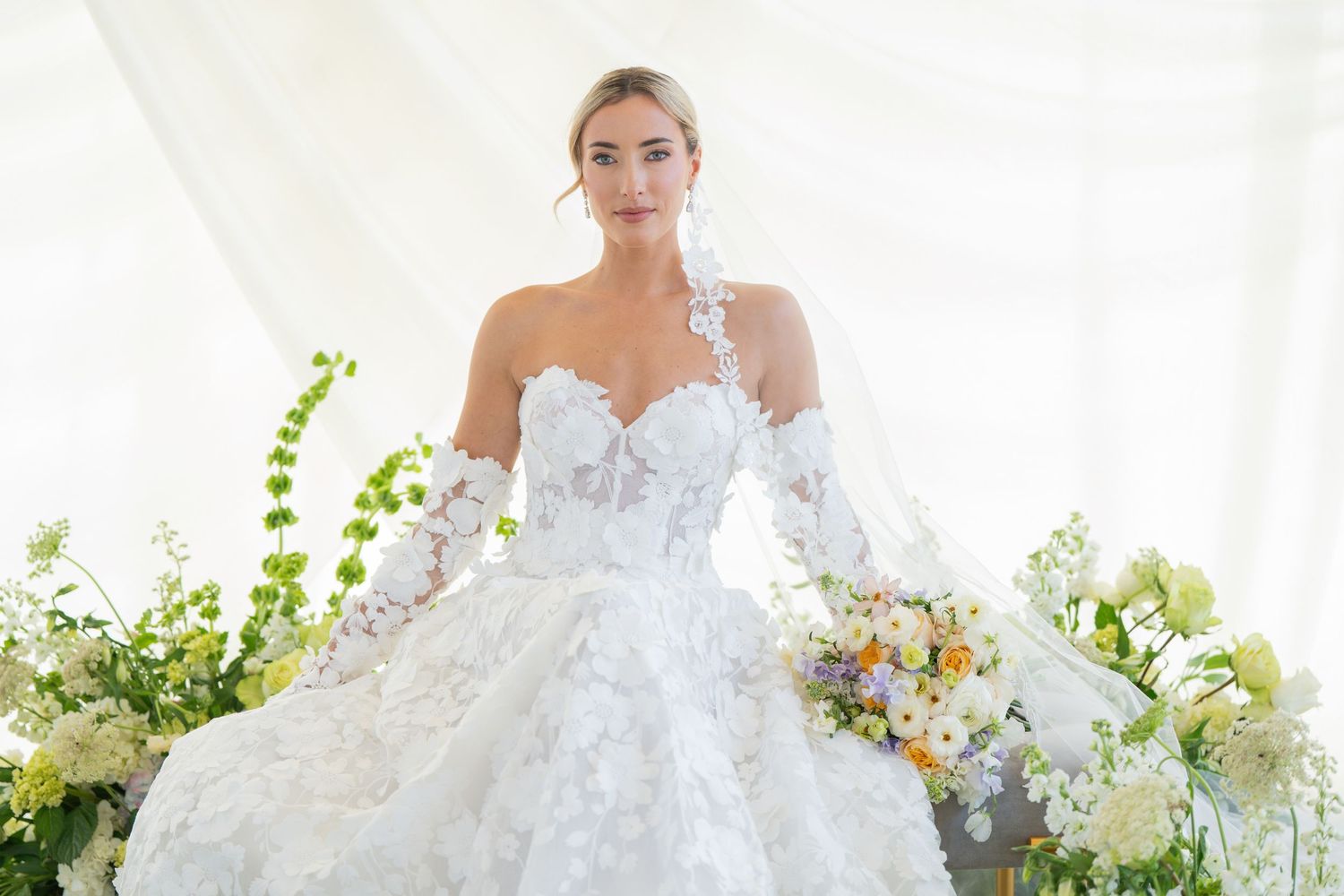Bride wearing one-shoulder lace wedding gown poses with spring bouquet among green foliage.