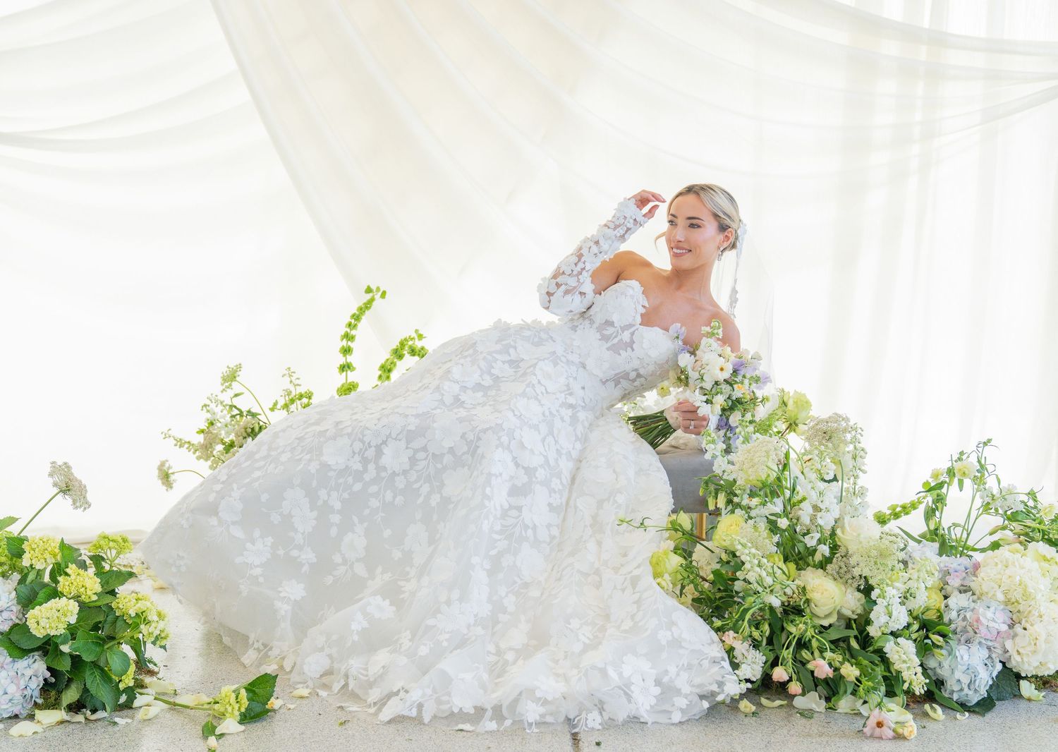 Ethereal bridal scene with flowing white dress surrounded by green and white flowers.