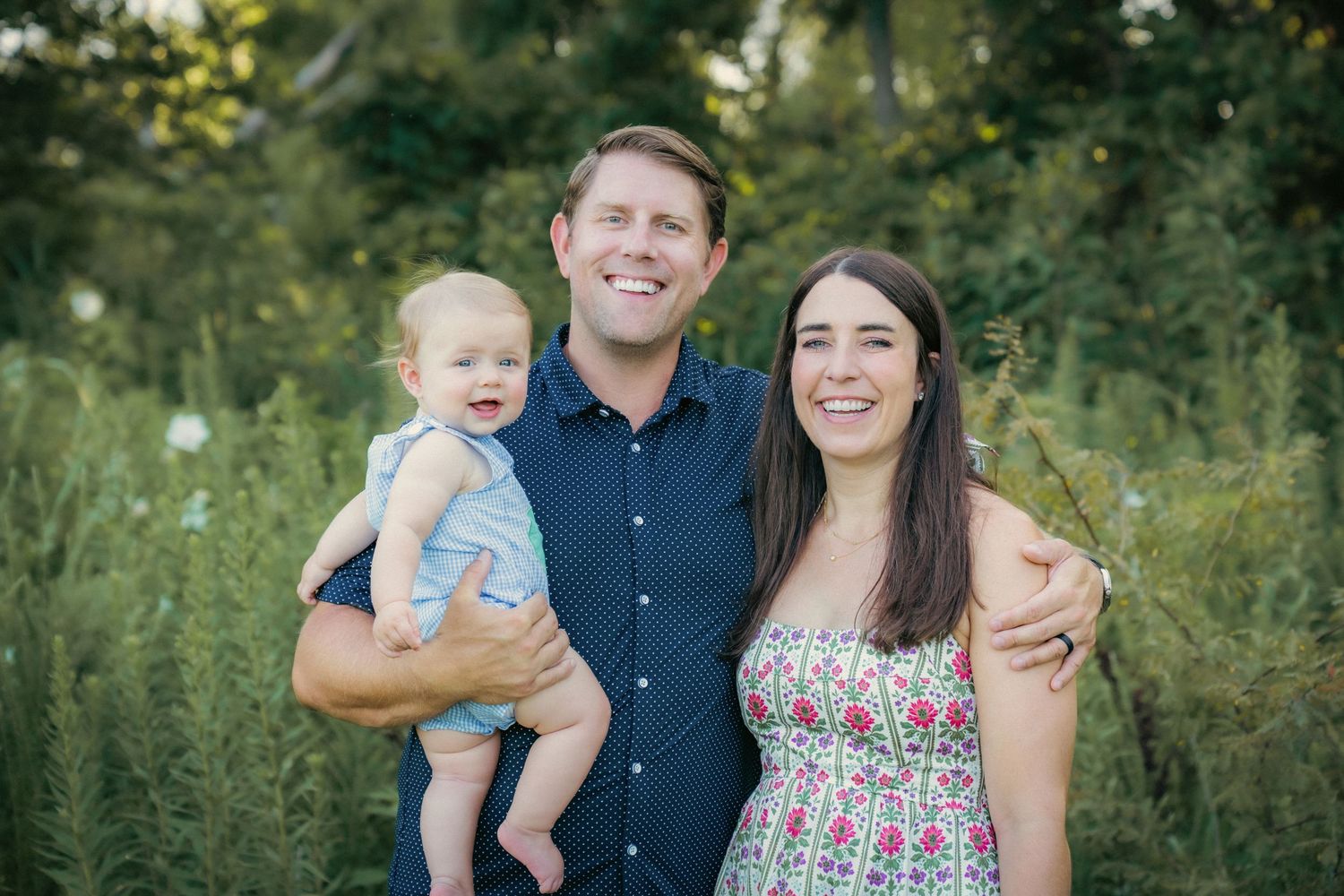 Family of three posing together outdoors with trees and greenery in background.
