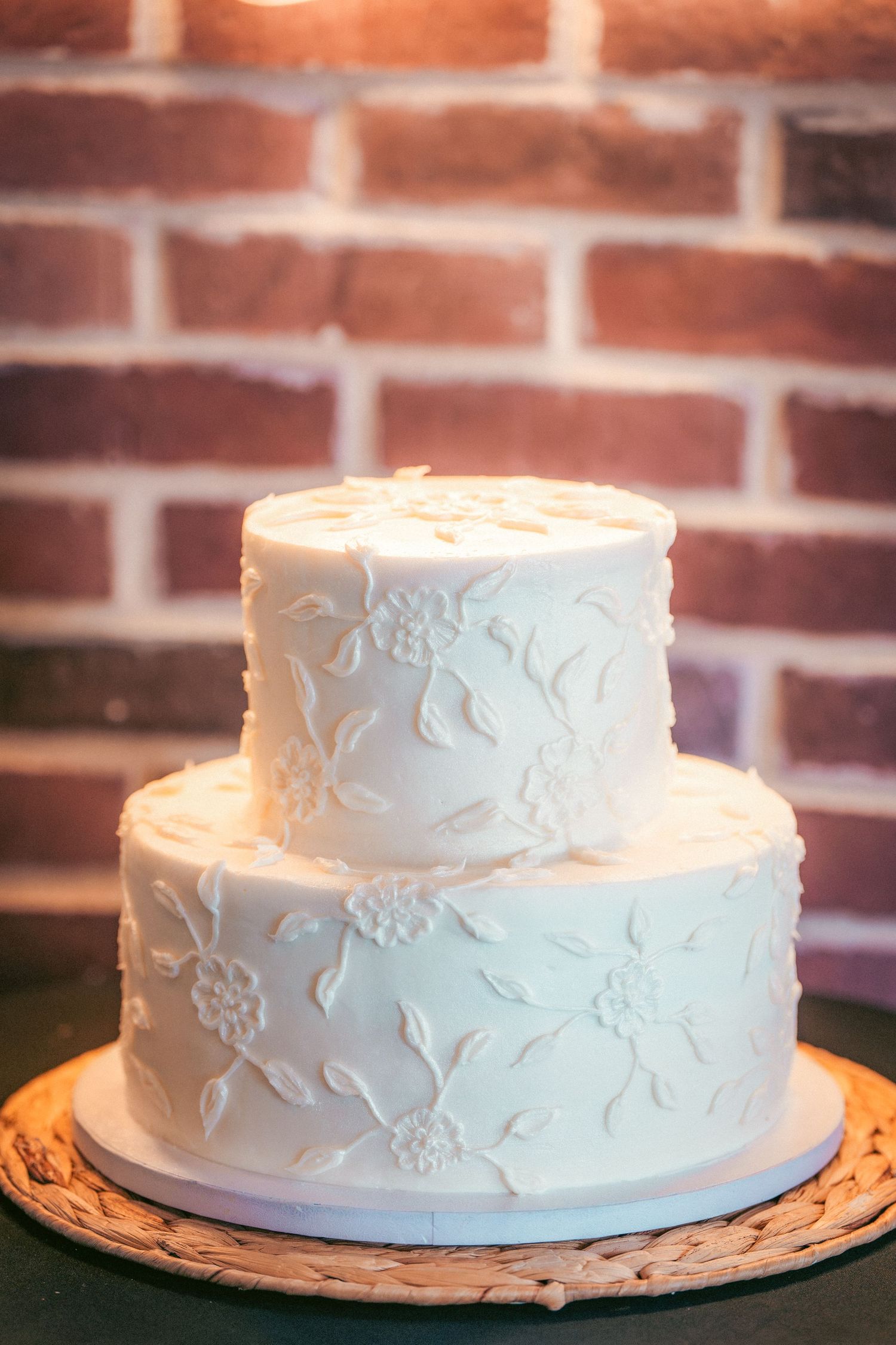 Two-tiered white wedding cake with elegant floral embossing against a brick wall background.
