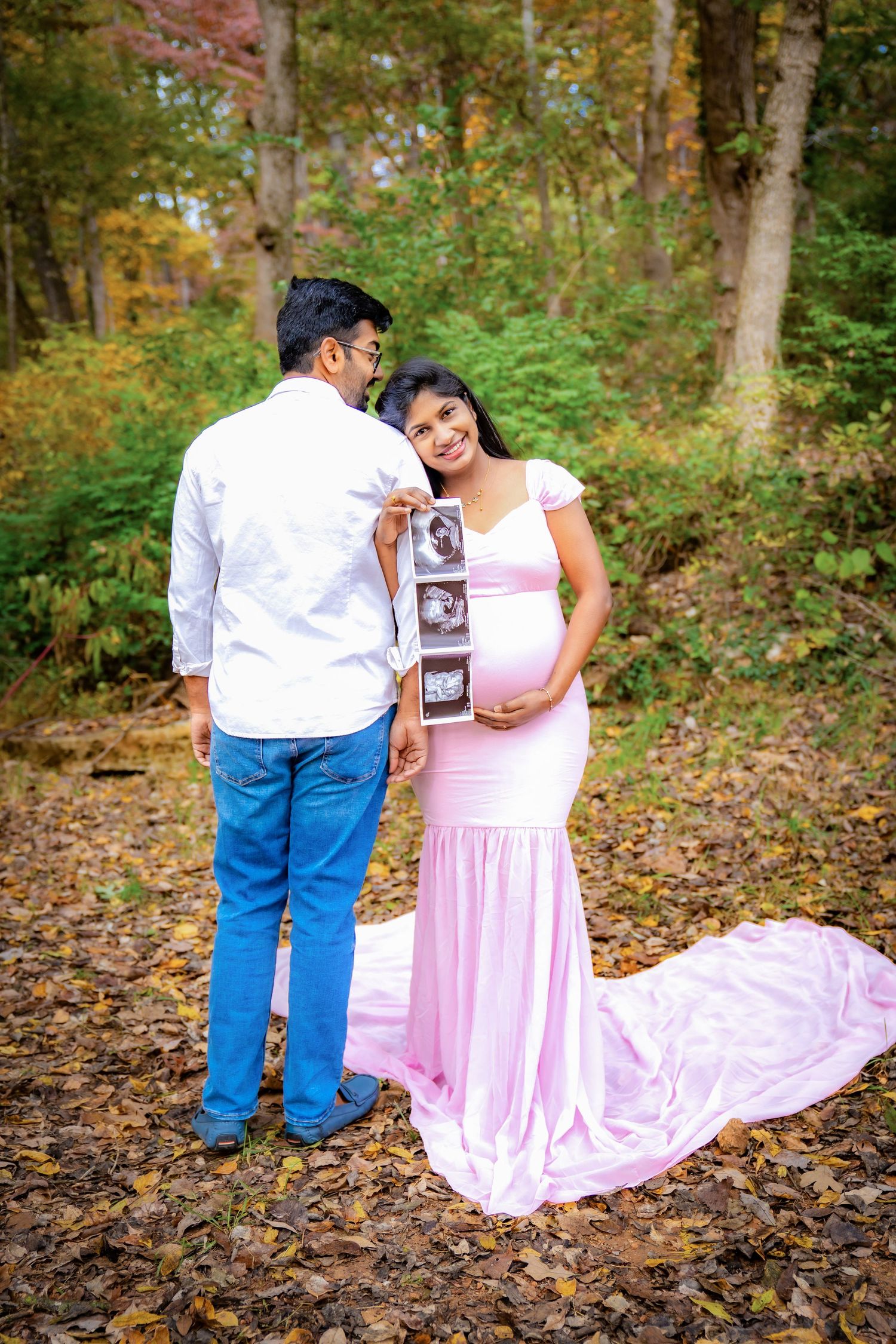 A couple holds ultrasound photos while posing in a wooded autumn setting.