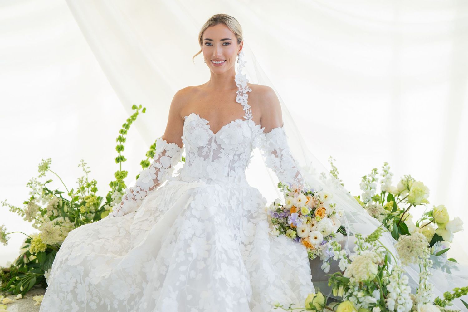 Bride in off-shoulder lace gown poses with floral bouquet against white drapery.