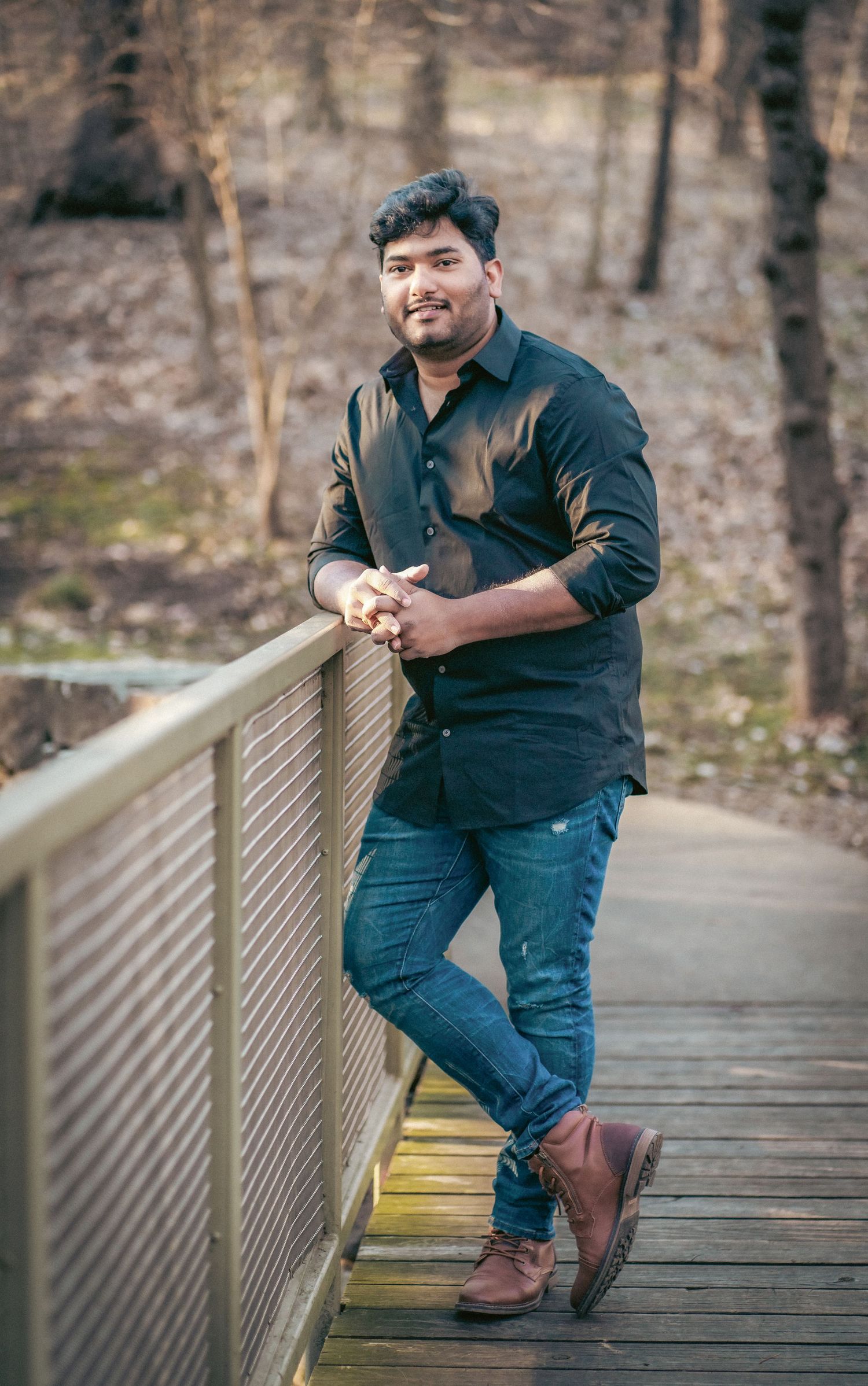 A person in dark clothing poses on a wooden bridge railing in a forest setting.