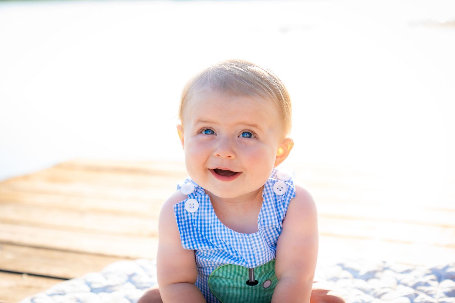 Baby in blue gingham outfit sitting on white blanket in bright natural light.