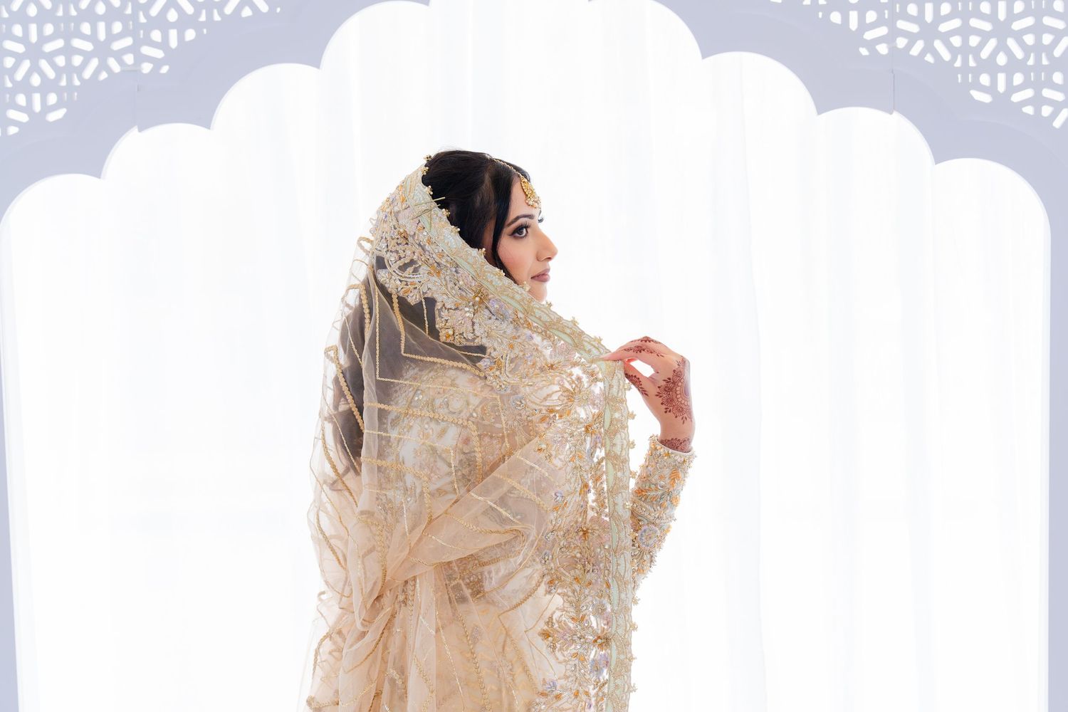 Traditional South Asian bride in ornate cream and gold outfit against white patterned backdrop.