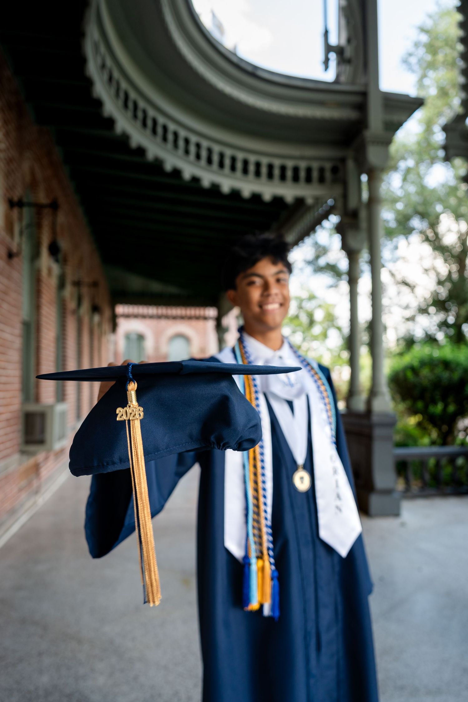 Smiling graduate in navy cap and gown holds mortarboard toward camera outside historic brick building.