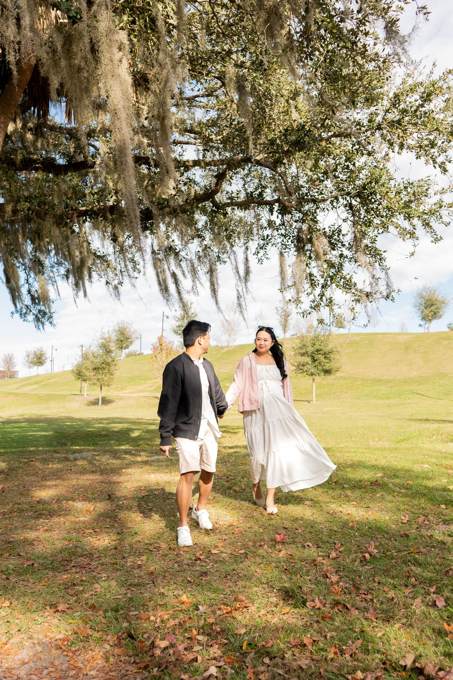 Couple walking hand in hand under Spanish moss-draped oak tree in a sunlit park.