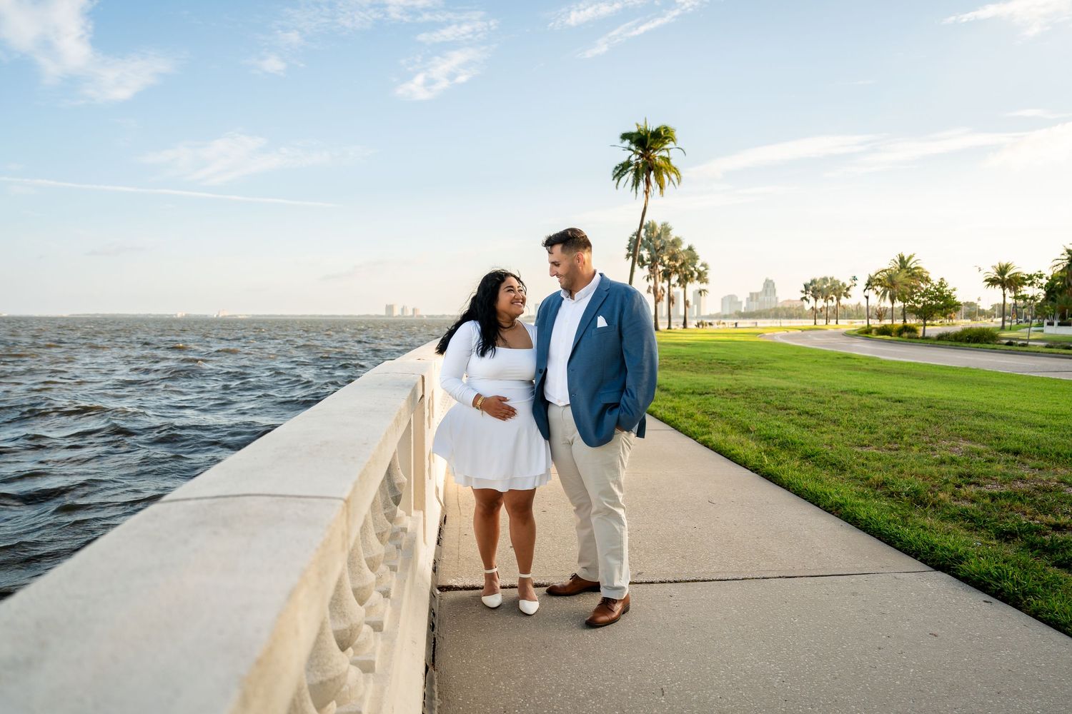 Couple posing on waterfront promenade with palm trees and city skyline, woman in white dress holding baby bump.