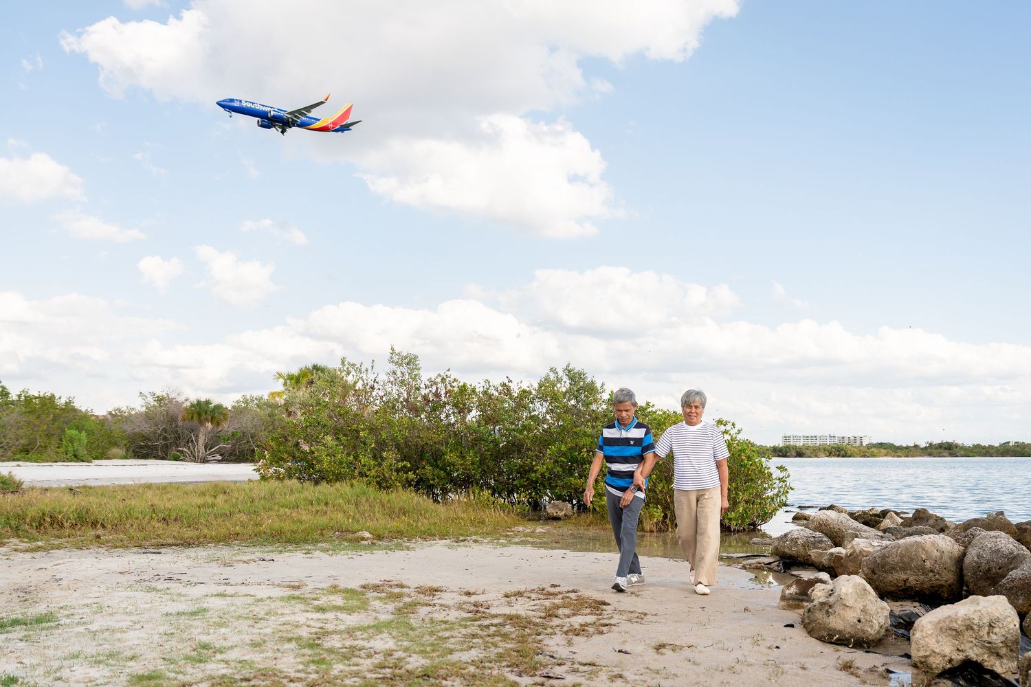 A man and boy walk along a rocky shoreline as a Southwest Airlines plane flies overhead.