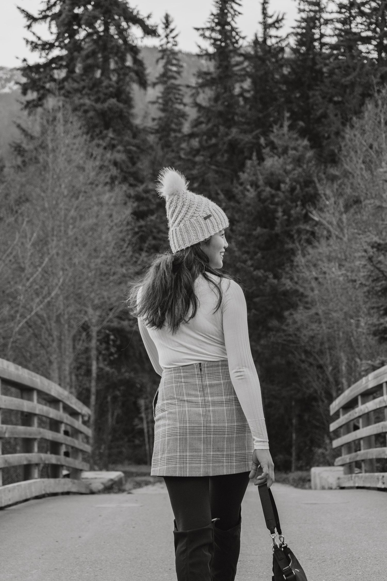 Young woman in plaid skirt and white turtleneck with pom beanie stands on bridge near forest.