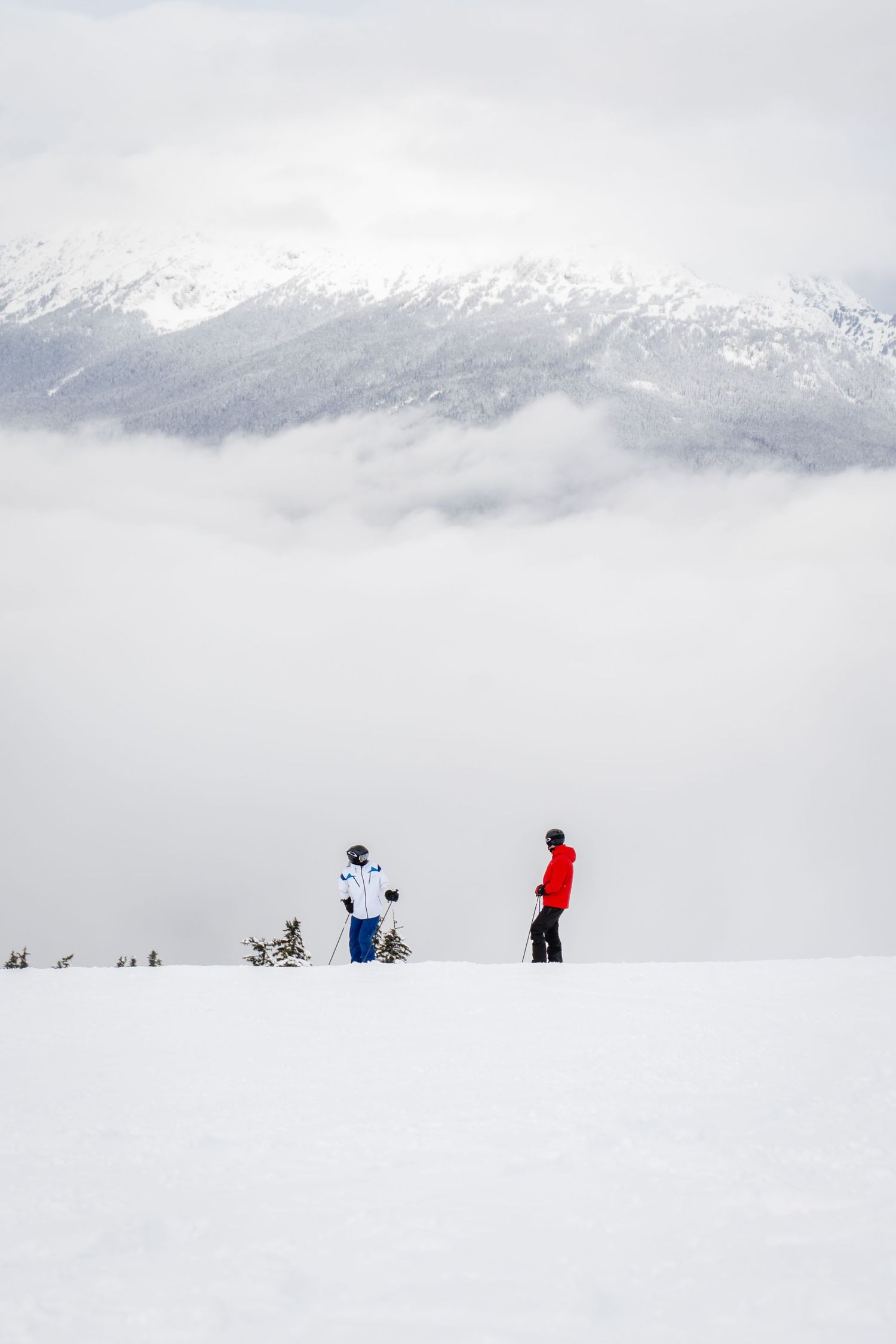 Two snowboarders standing on snowy mountain ridge with dramatic cloud-covered peaks in background.