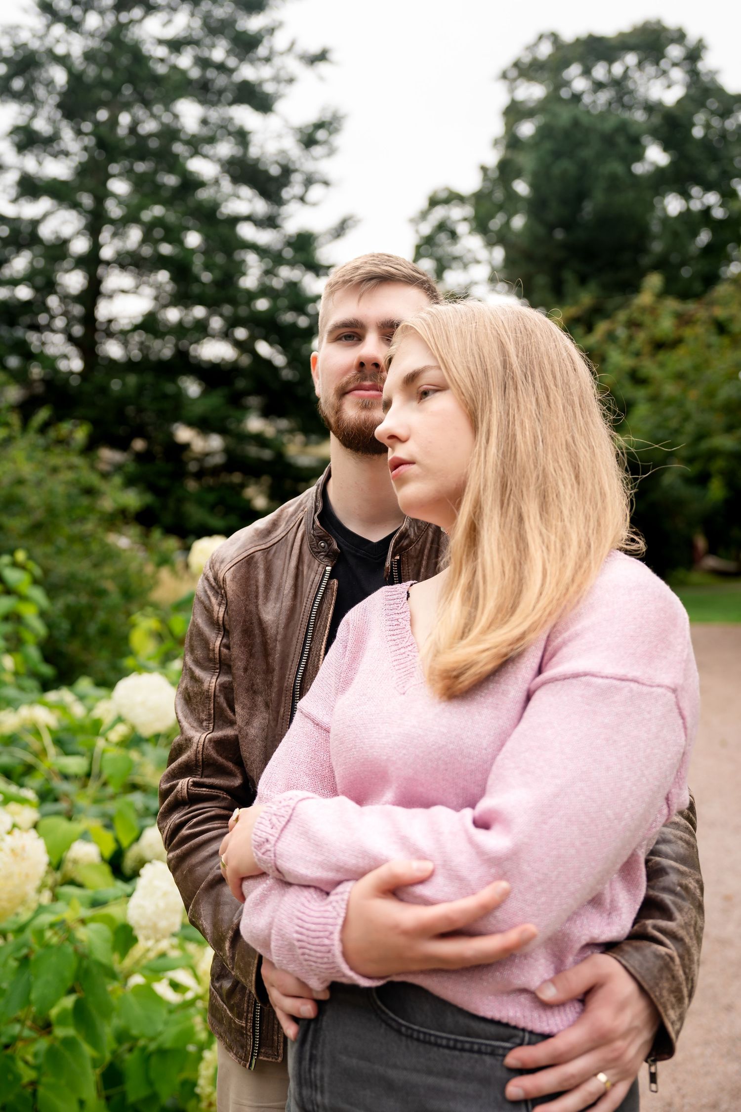 A couple embraces in a lush green park, the man holding the blonde woman in a pink sweater from behind.