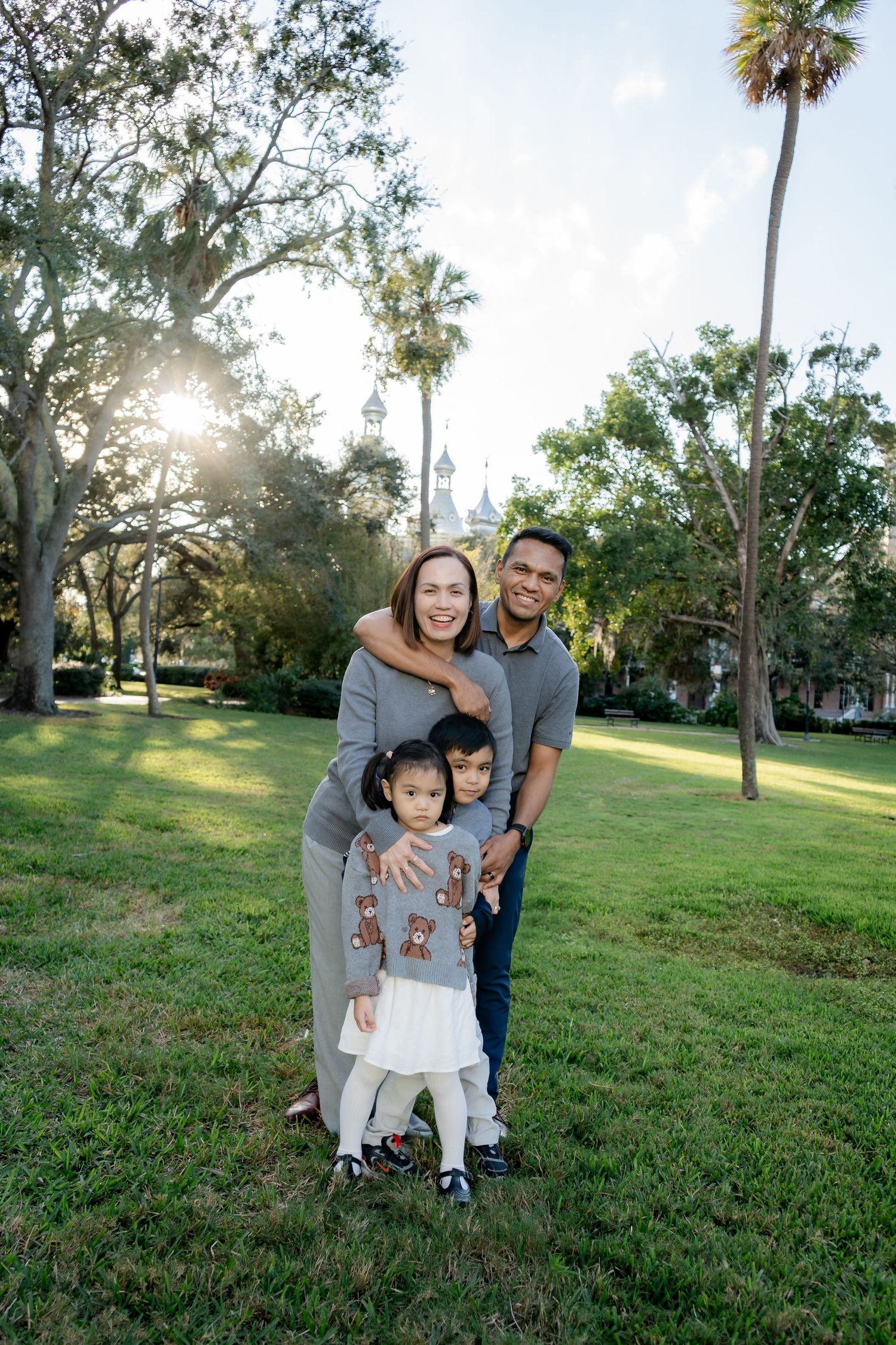 A happy family of four poses together on a sunny green lawn with palm trees and a historic building behind them.