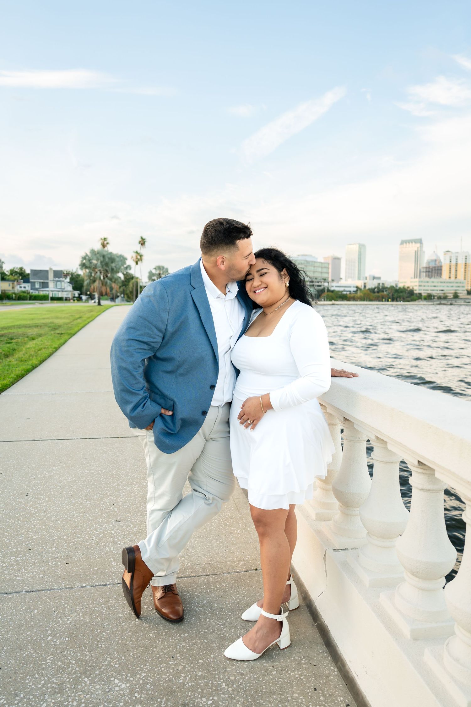 Couple sharing a romantic moment by a waterfront promenade, man in blue blazer kissing woman in white dress.