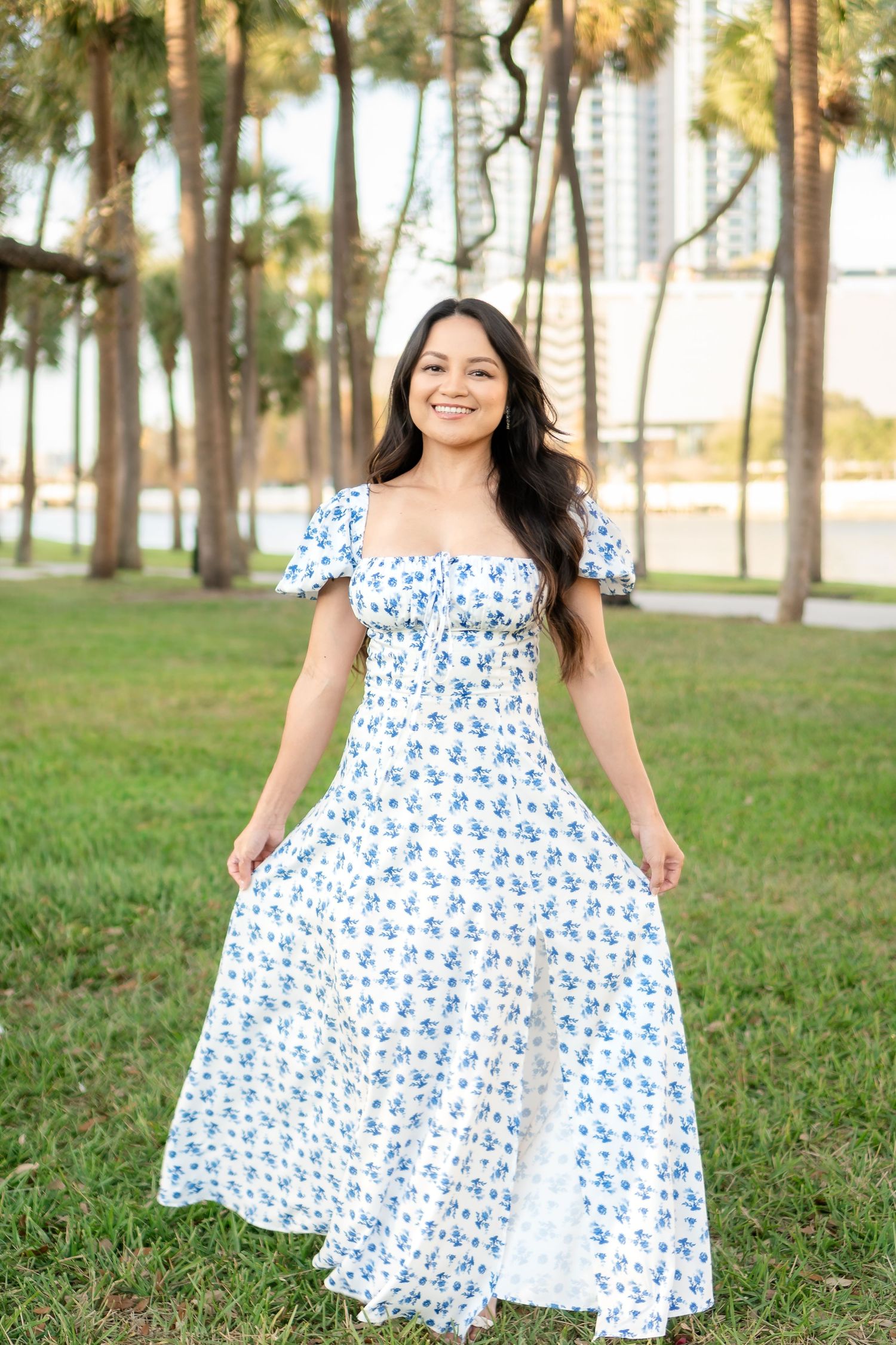 Woman twirling in a white and blue floral maxi dress in a park with palm trees.