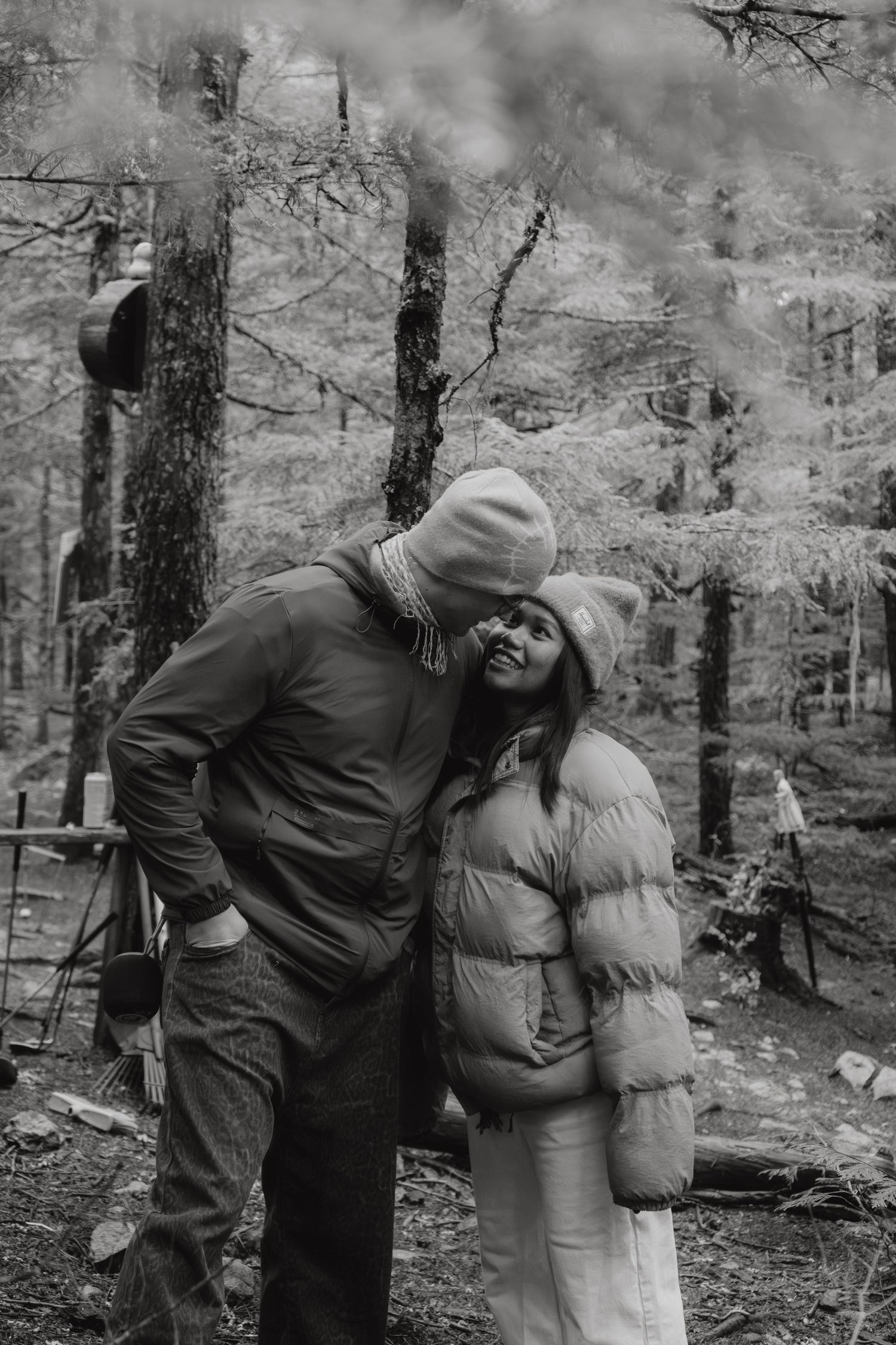 A black and white photo of a couple embracing warmly in a dense forest wearing winter jackets.