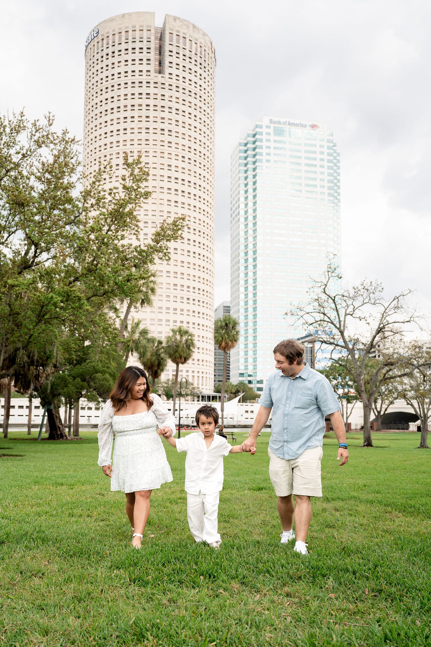 A family of three walks hand-in-hand on a grassy park lawn with city skyscrapers in the background.