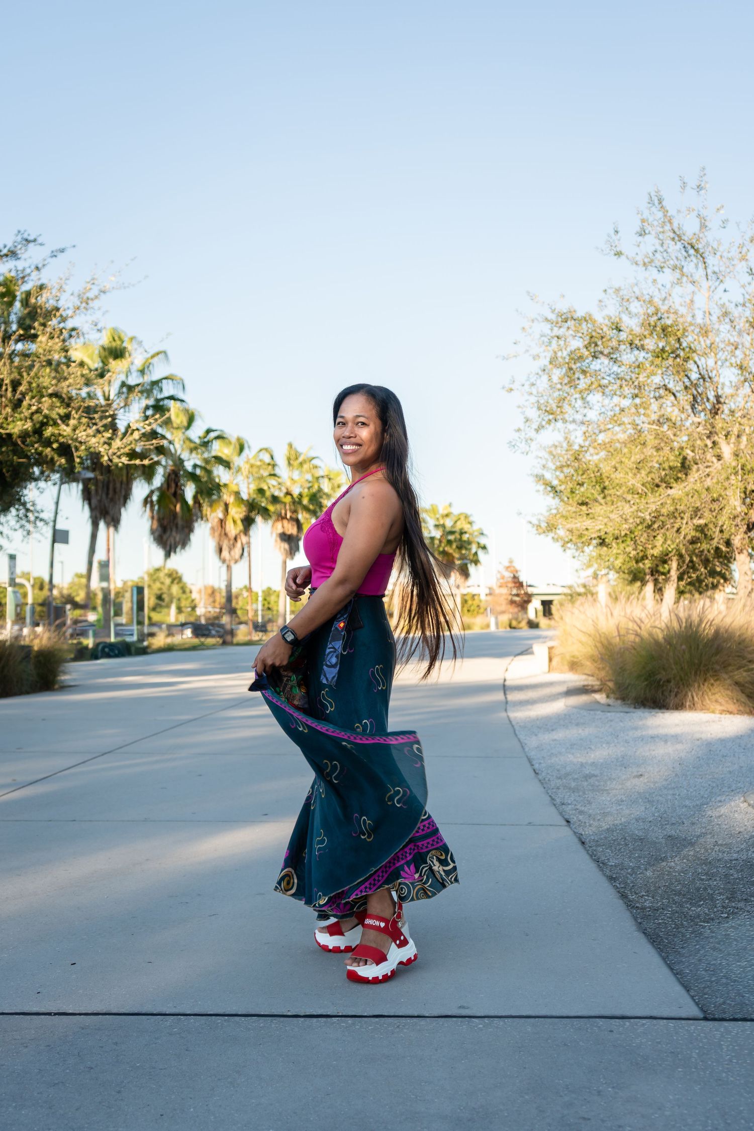 Smiling woman in pink top and patterned maxi skirt posing on a palm tree-lined walkway.