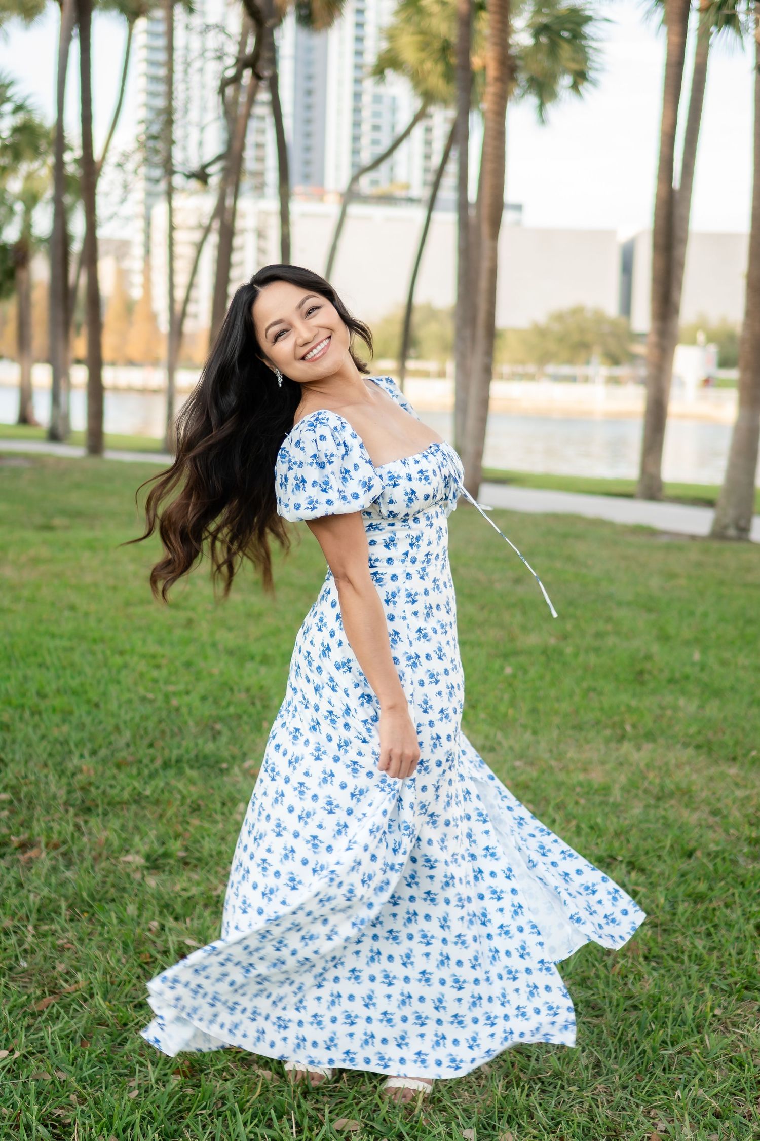 Woman in a white floral maxi dress spinning joyfully in a park with palm trees and a waterfront.