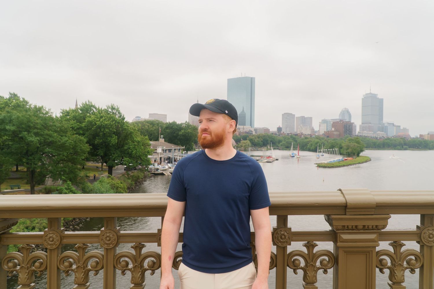 Bearded man in navy shirt leaning on ornate bridge railing with Boston skyline and river behind him.