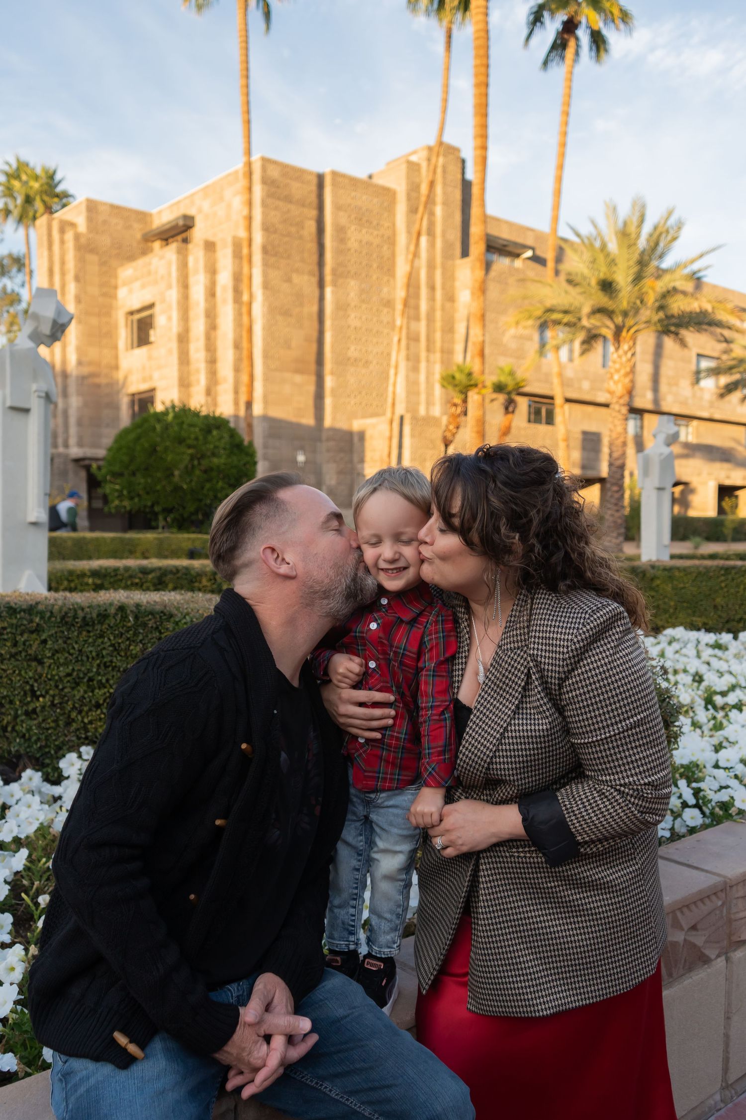 Parents kissing smiling toddler boy on cheeks outdoors with palm trees and building in background.