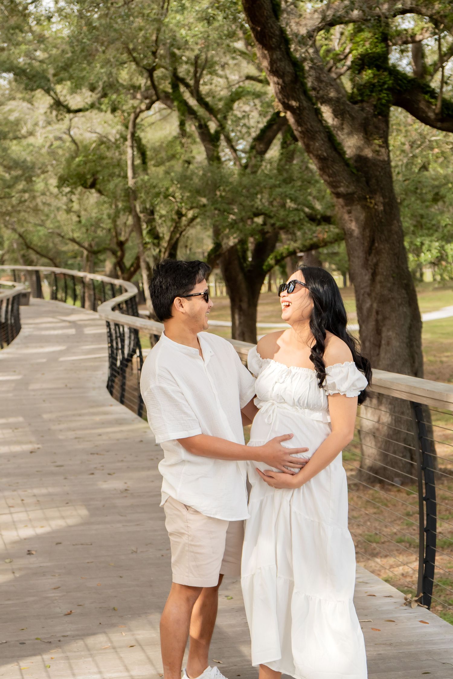 Happy pregnant couple in white outfits embracing on wooden bridge surrounded by lush green trees.