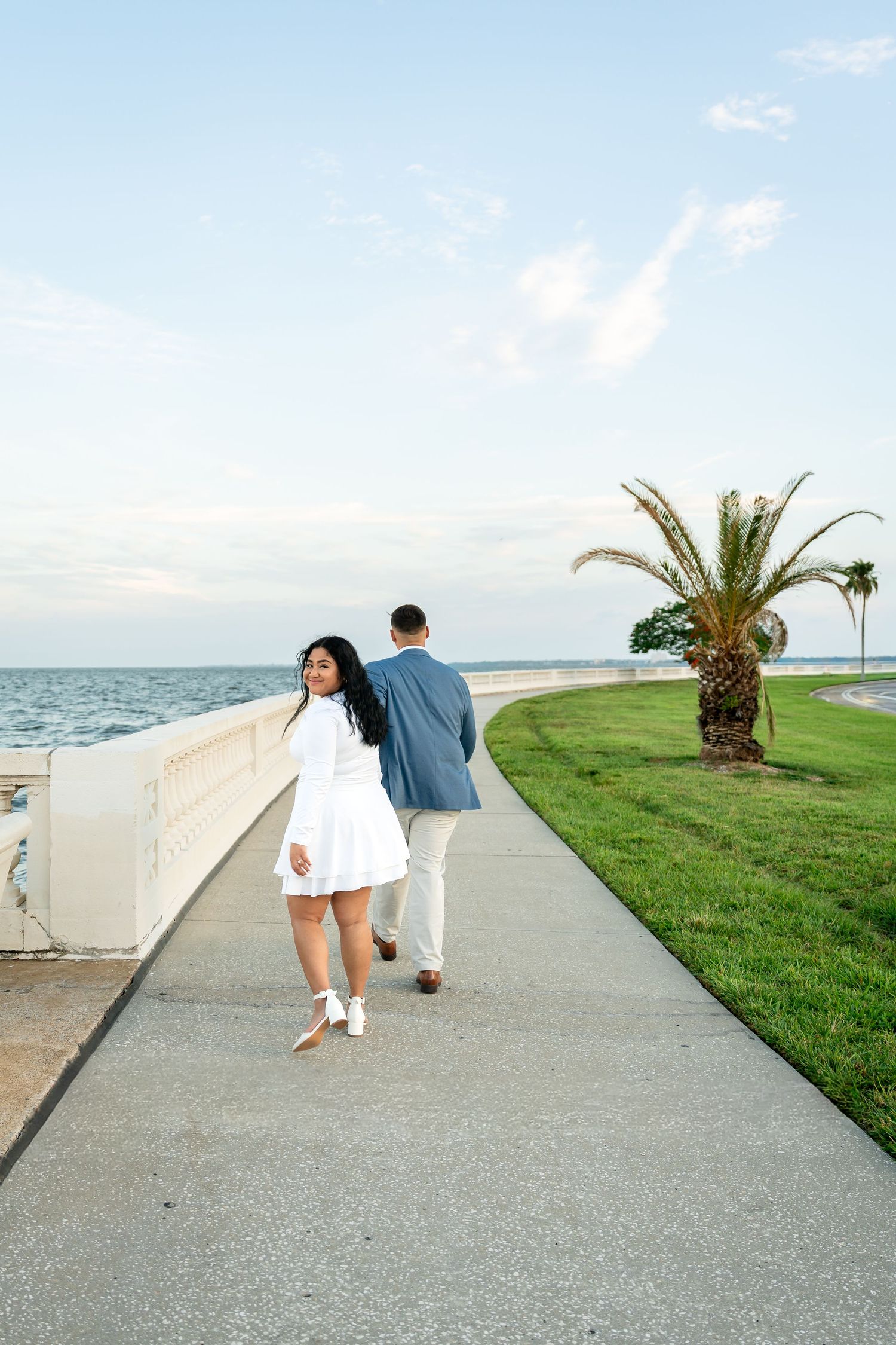 Couple walking along a waterfront promenade, woman in white dress glancing back, man in blue blazer, palm tree and ocean views.