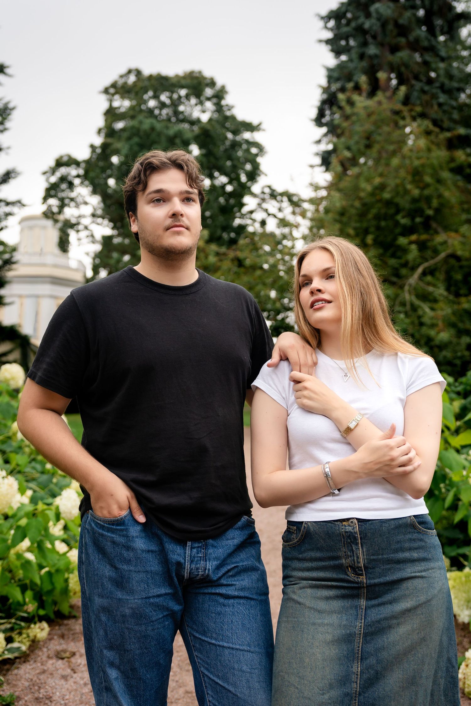 A young couple poses in a lush garden, man in black tee and jeans, woman in white tee and denim skirt.