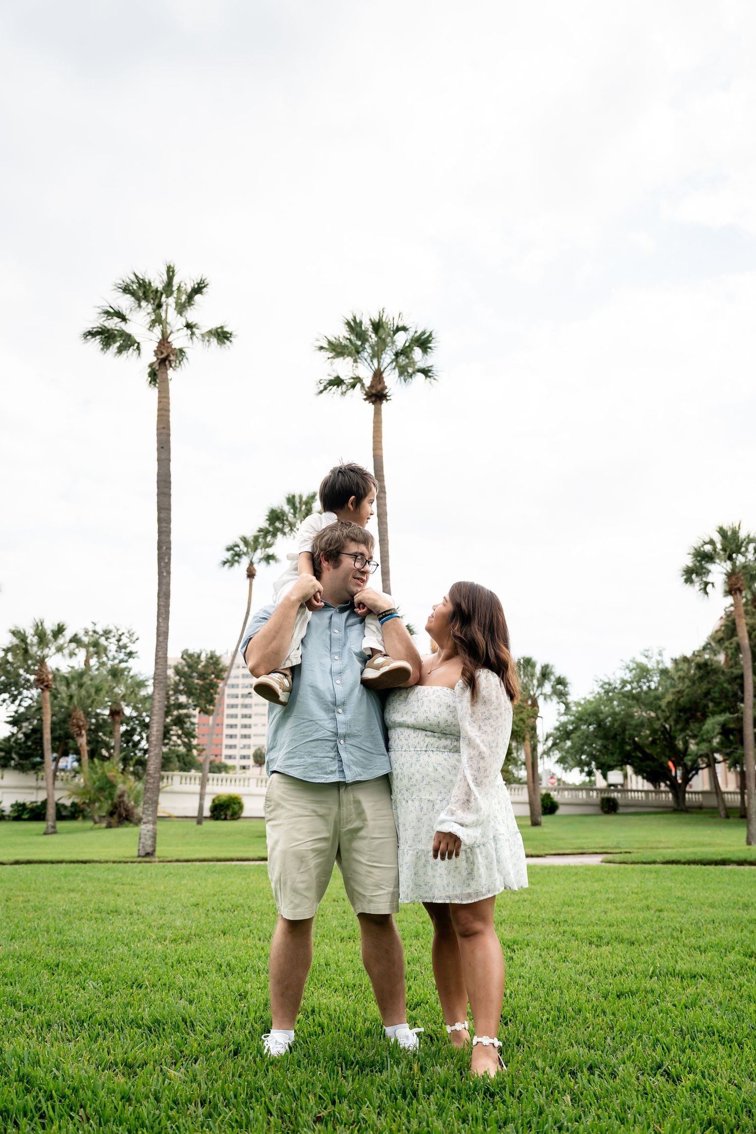 A family of three smiles together on a green lawn with tall palm trees and a cloudy sky above.