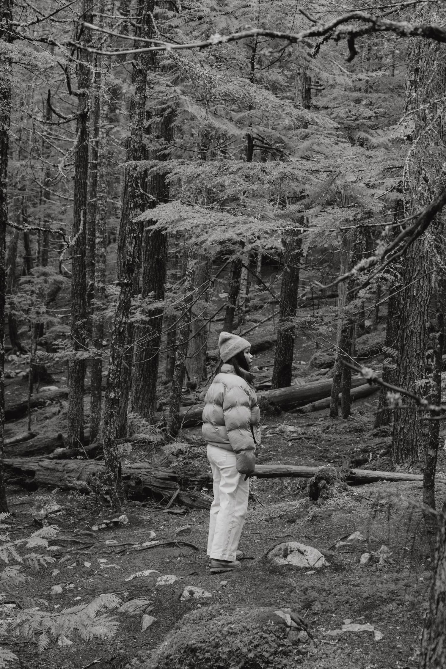 Black and white photo of a woman in a puffer jacket standing in a dense forest trail.