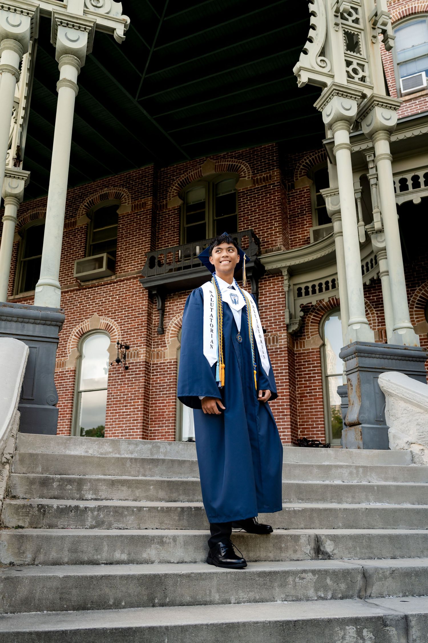 Graduate in navy blue cap and gown stands proudly on stone steps before ornate brick building.