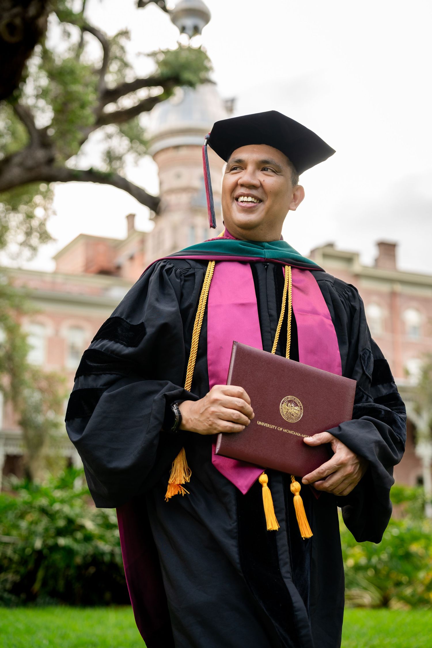 Smiling graduate in black doctoral gown, green hood, and cap holds diploma outdoors near historic brick building.