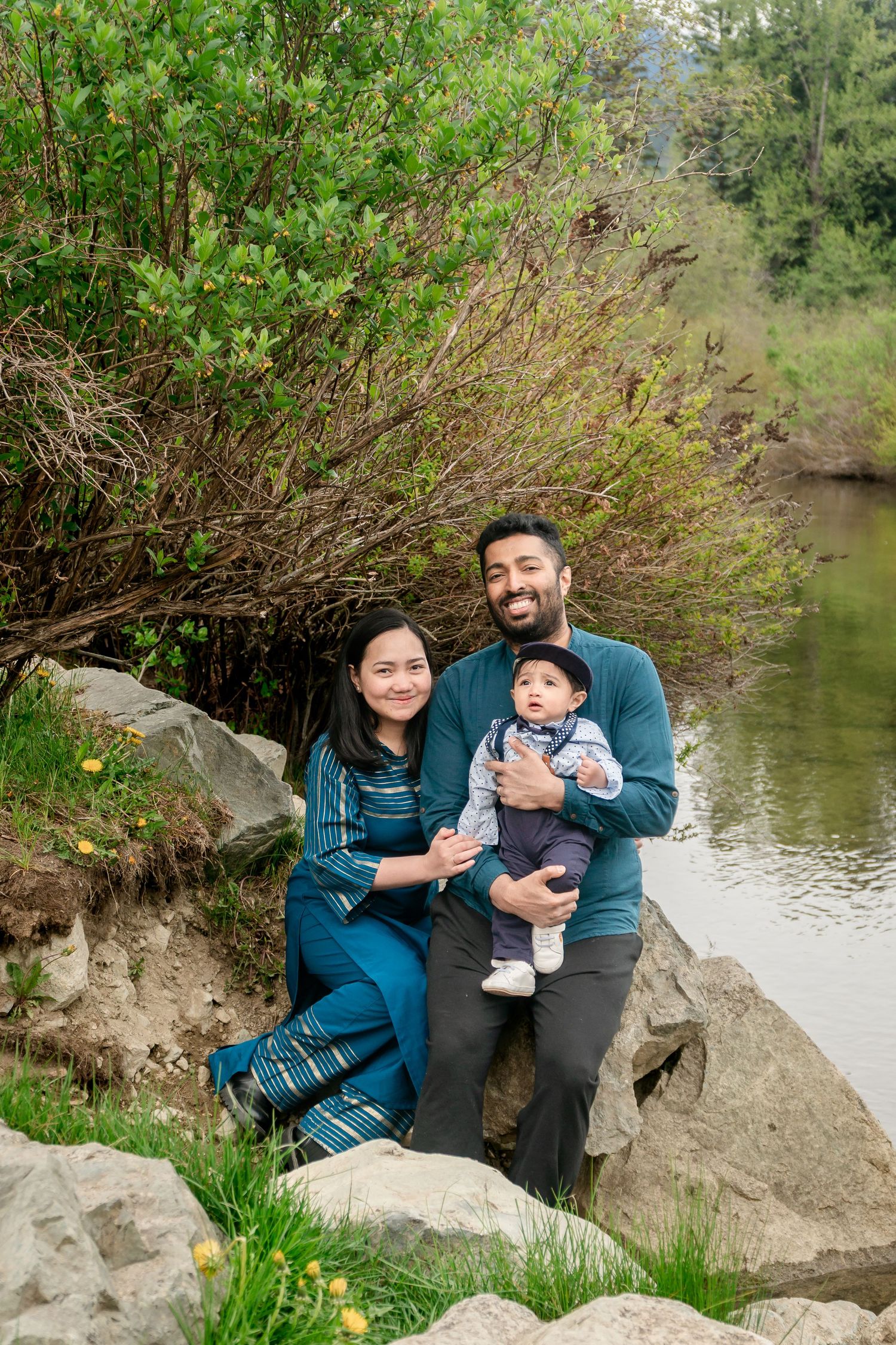 Happy family of three with infant sits on rocks beside a peaceful green creek.