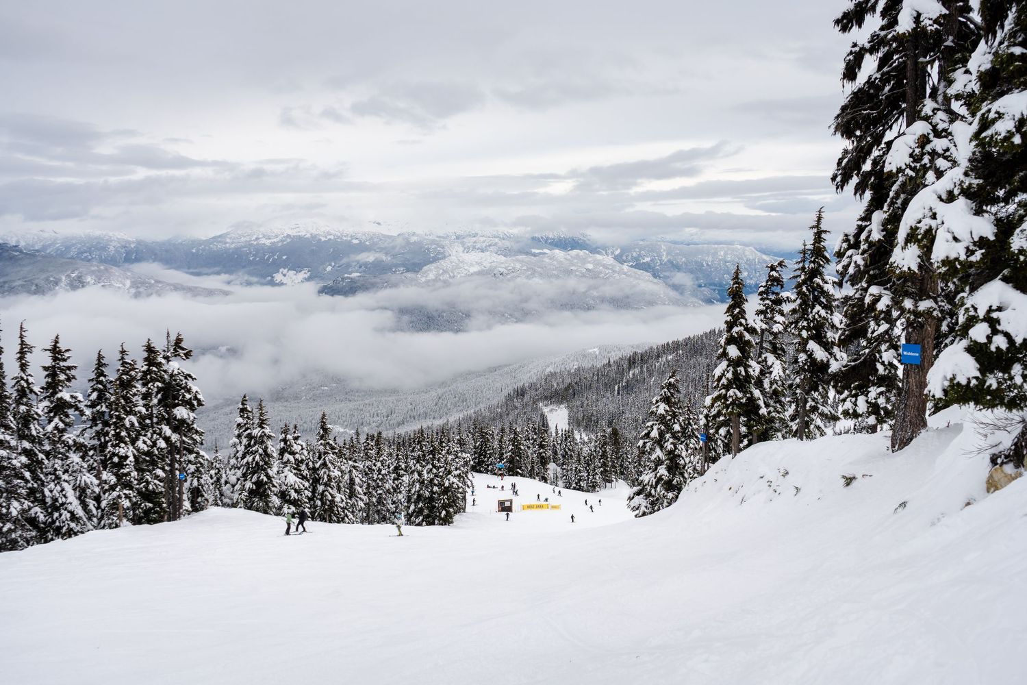 Snow-covered mountain ski resort with evergreen trees and clouds rolling over valley below.