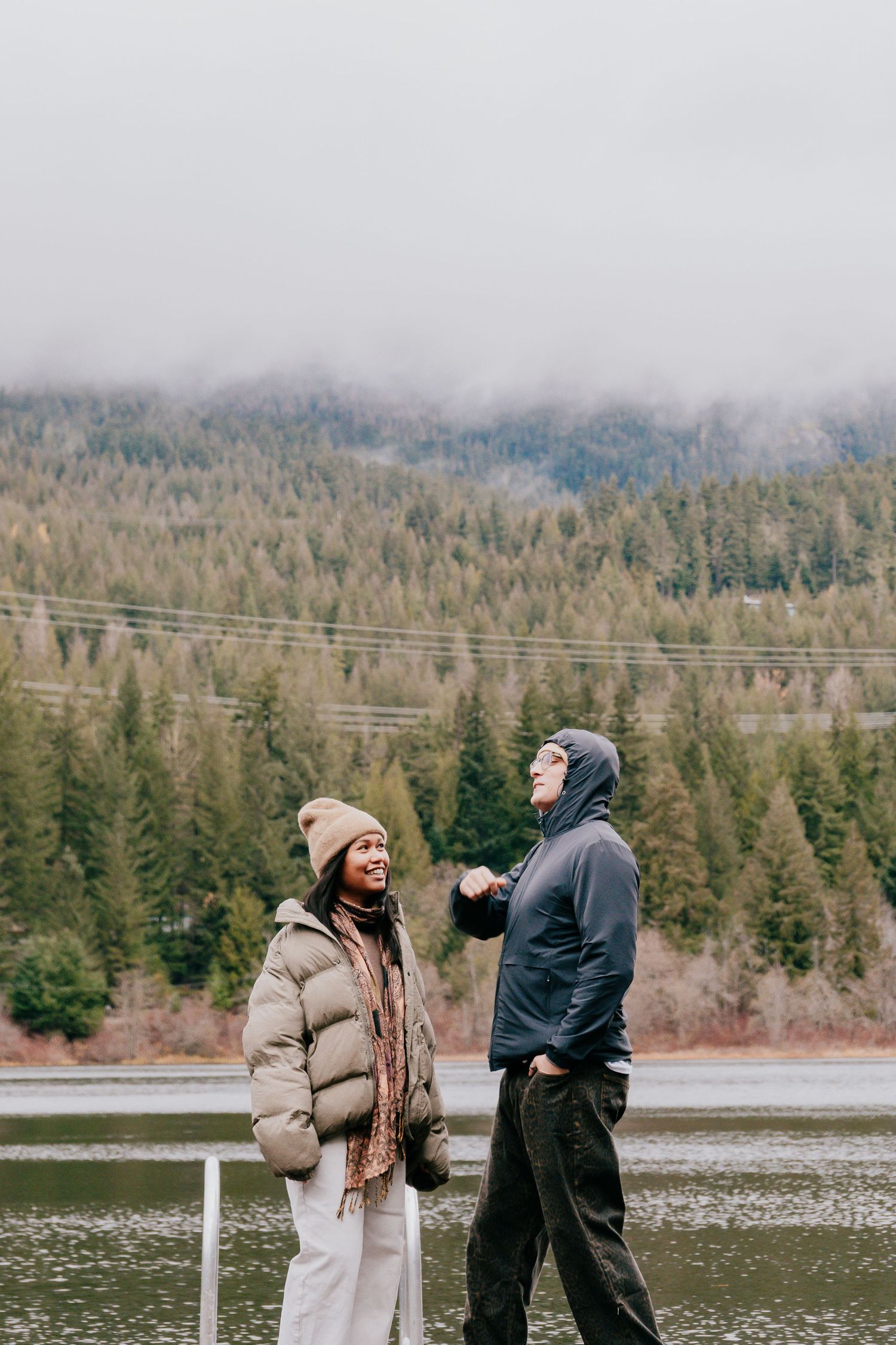 A smiling couple in winter jackets chatting by a misty lake with a forested mountain backdrop.