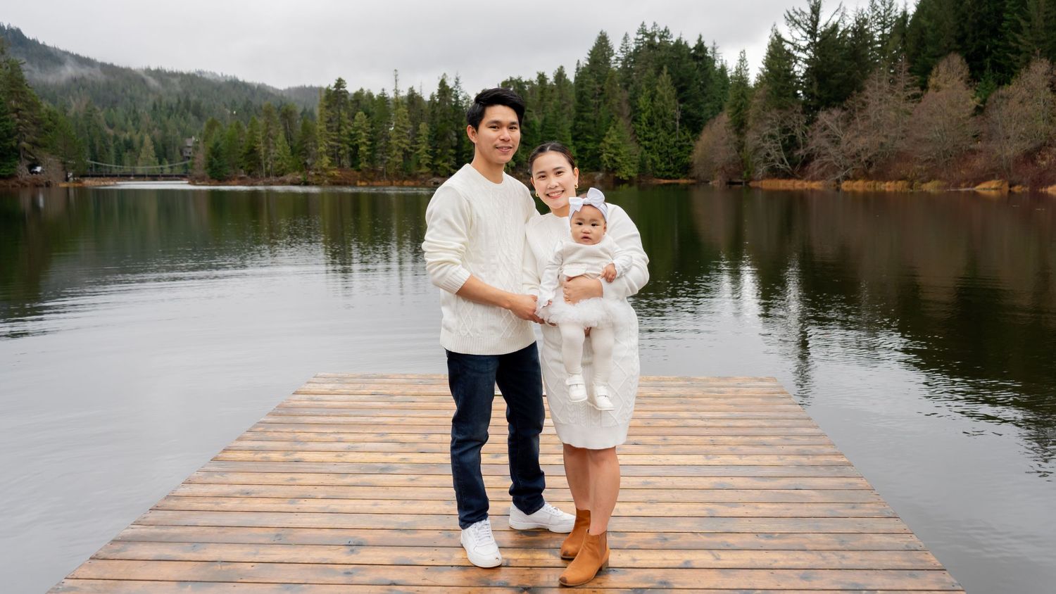 Young family with baby posing on wooden dock by serene mountain lake surrounded by evergreen forest.