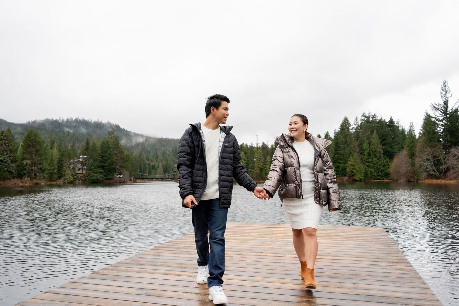 A couple holds hands and walks on a wooden dock beside a calm lake surrounded by evergreen trees.