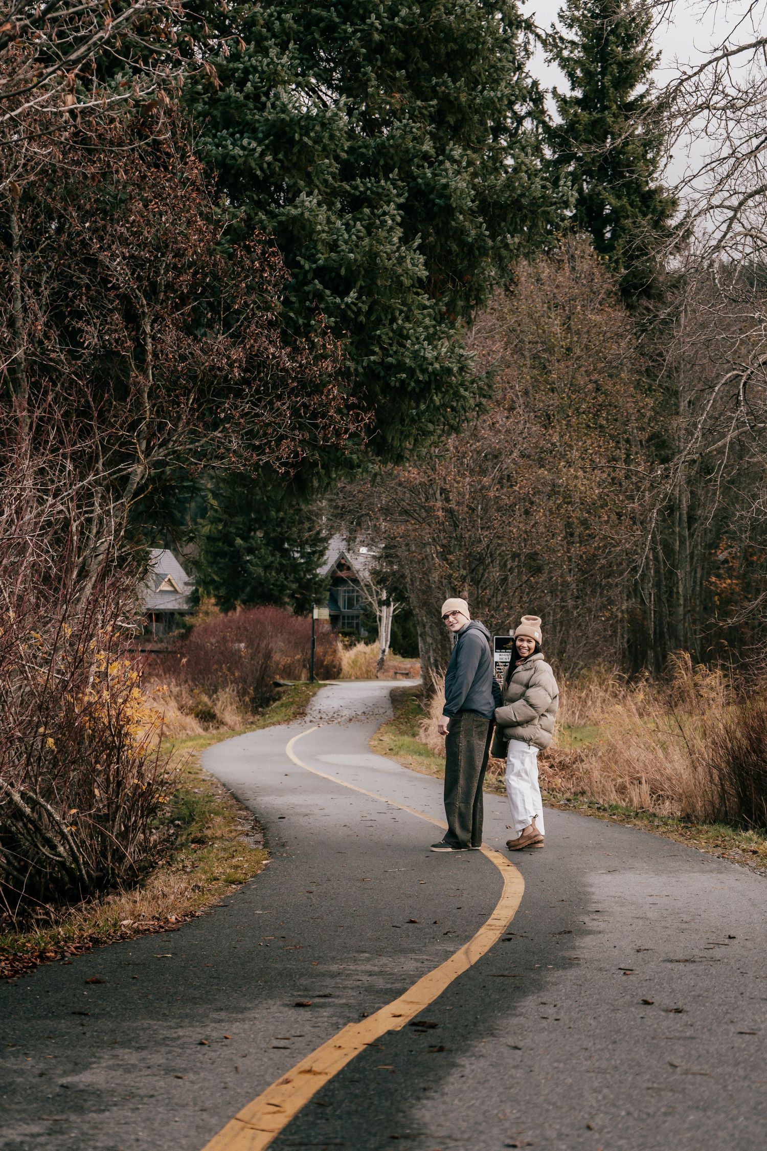 A couple holding hands and posing on a winding road lined with autumn trees and shrubs.