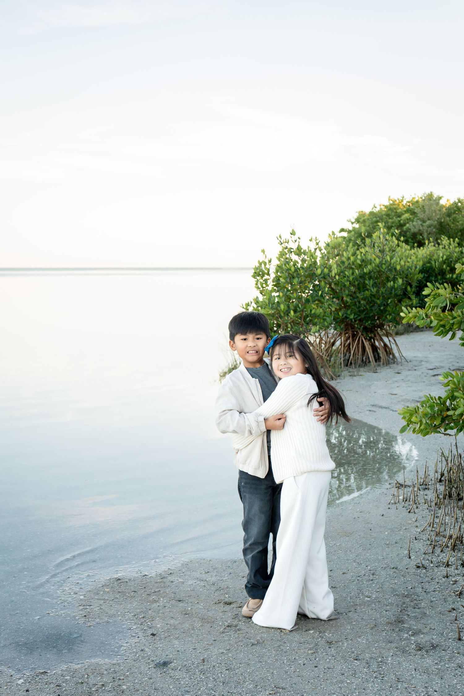 Two young children embrace and smile while standing barefoot on a sandy beach with mangroves behind them.