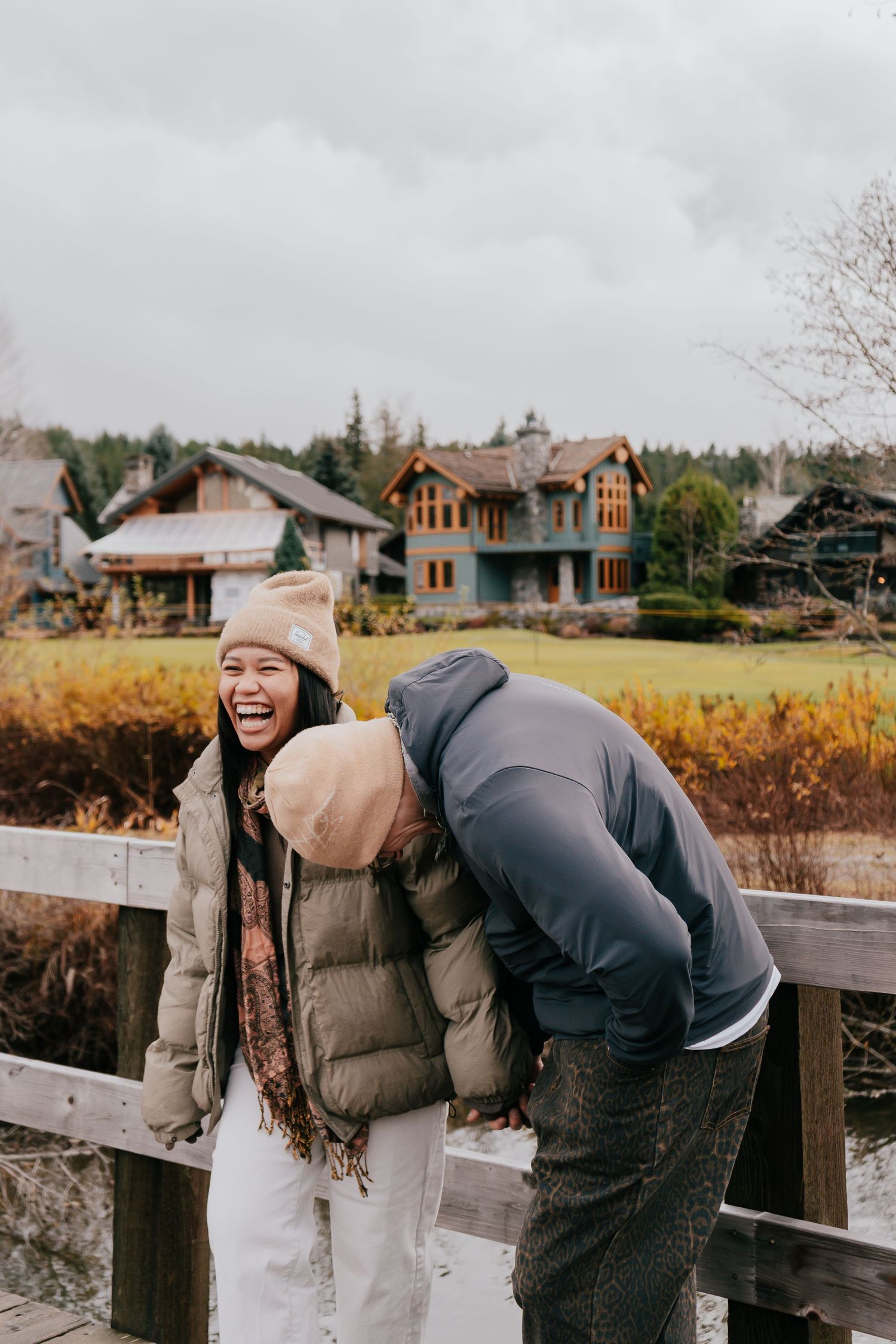 Laughing couple in winter jackets and beanies on wooden bridge with rustic house behind them.