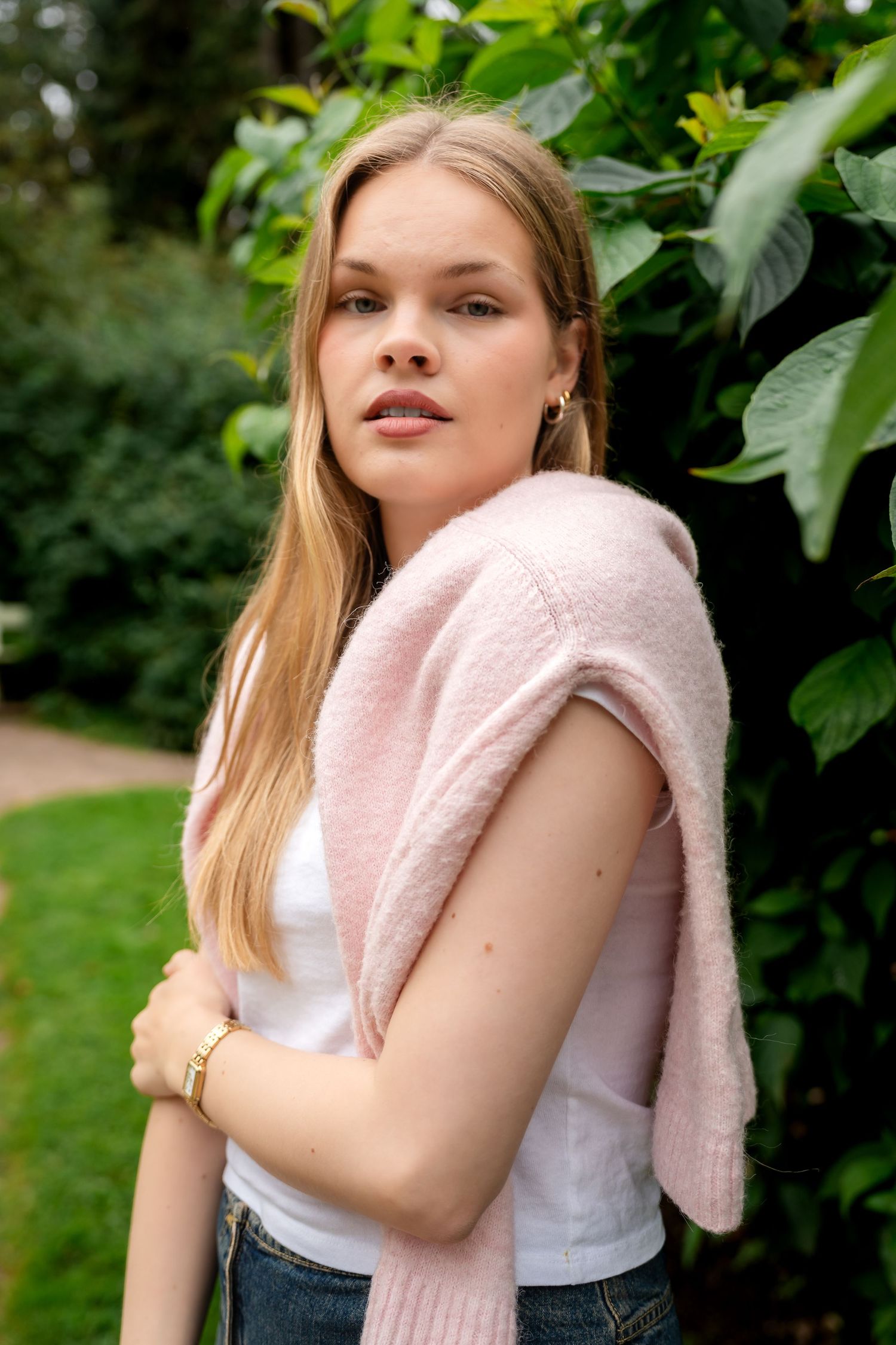 Young blonde woman in white tee with pink sweater over shoulders, posing outdoors near green foliage.