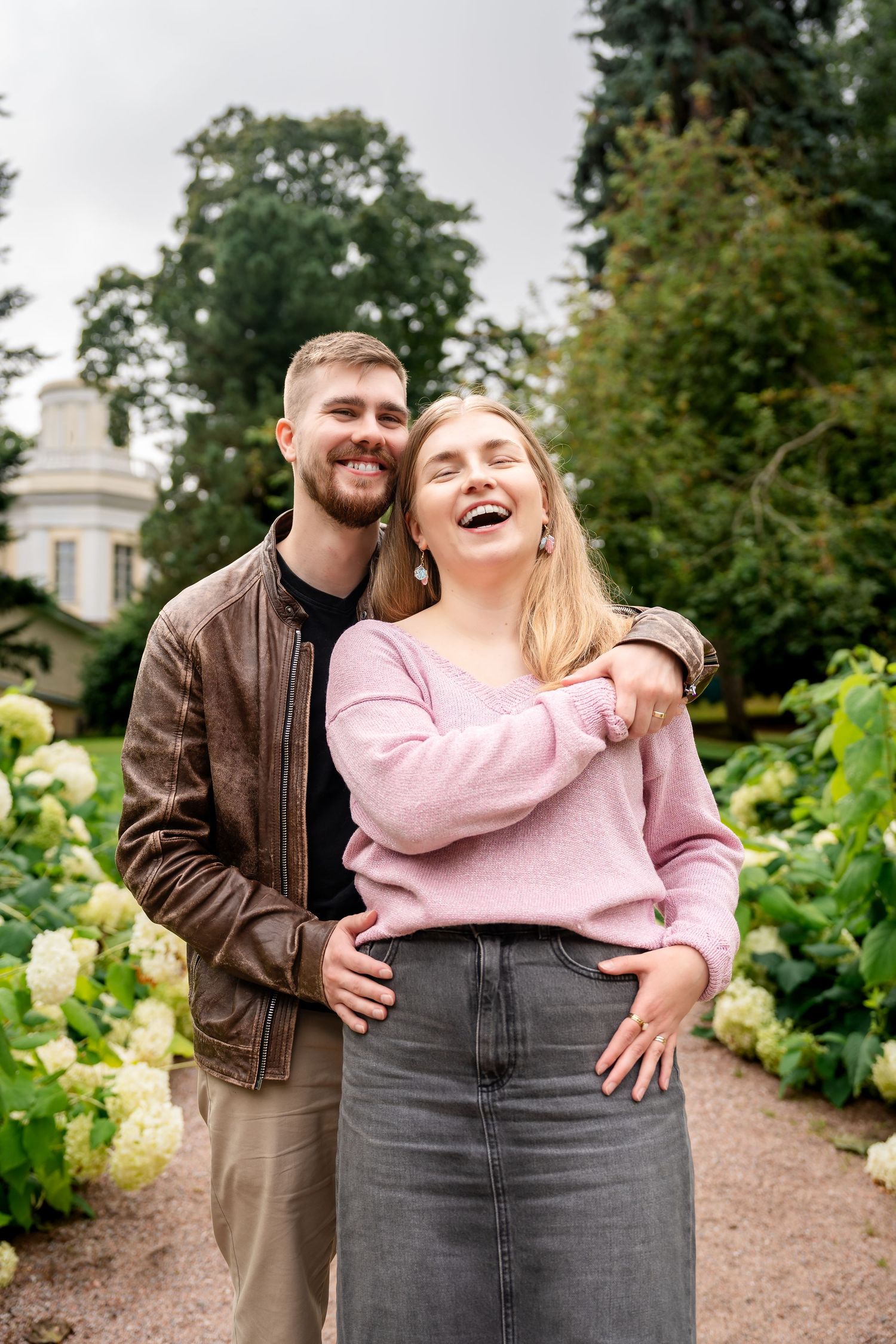 A happy couple embraces in a lush garden, woman in pink sweater laughing joyfully, man in brown leather jacket smiling.