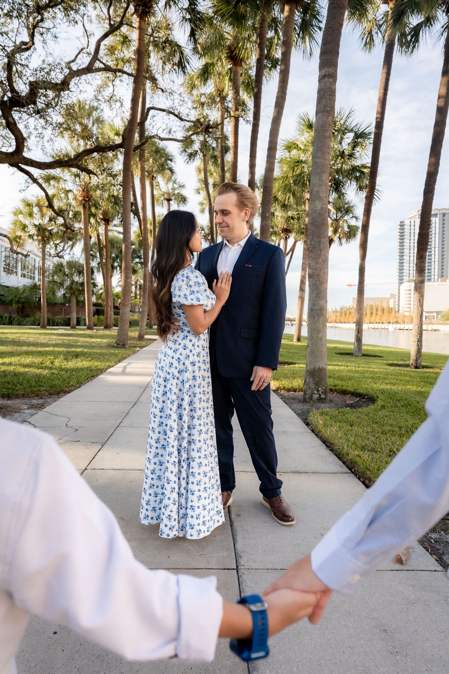 Young couple in elegant attire smiling at each other on a palm-lined sidewalk at golden hour.