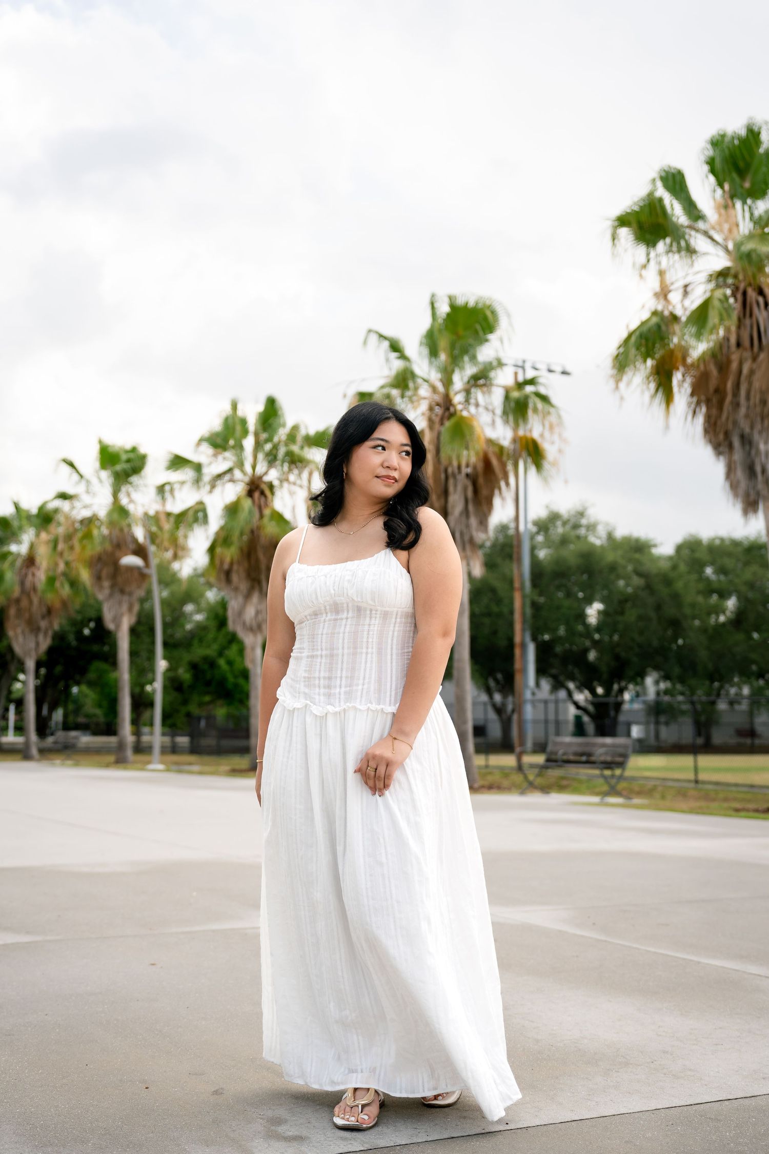 Woman in white maxi dress standing outdoors with palm trees in the background on a cloudy day.