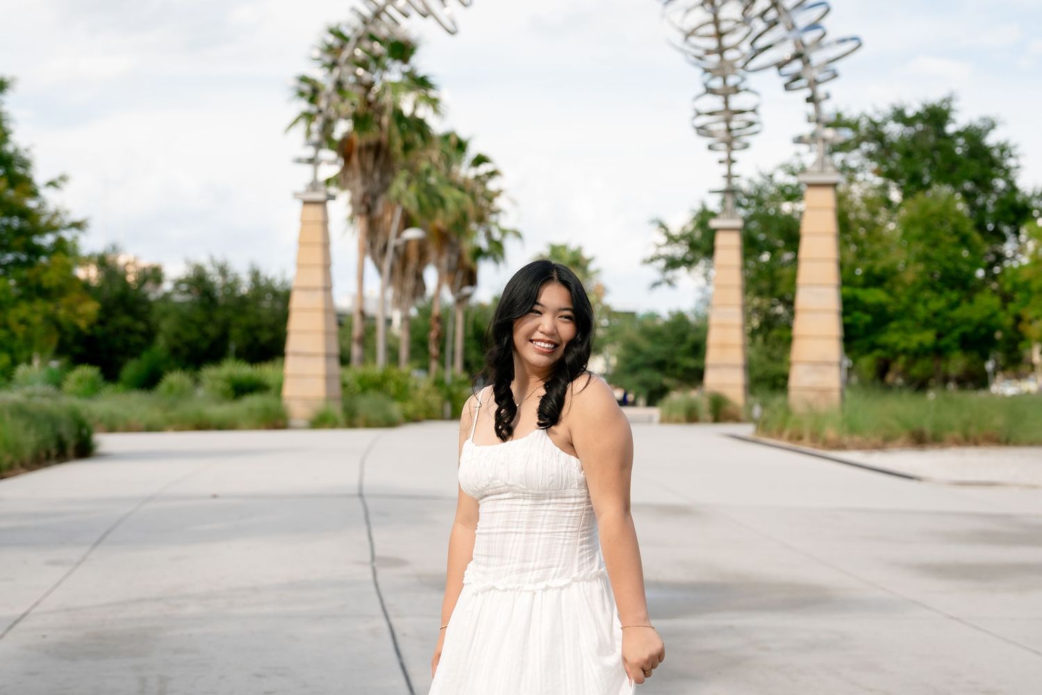 Young woman in white dress smiling outdoors near palm trees and decorative columns in a park.