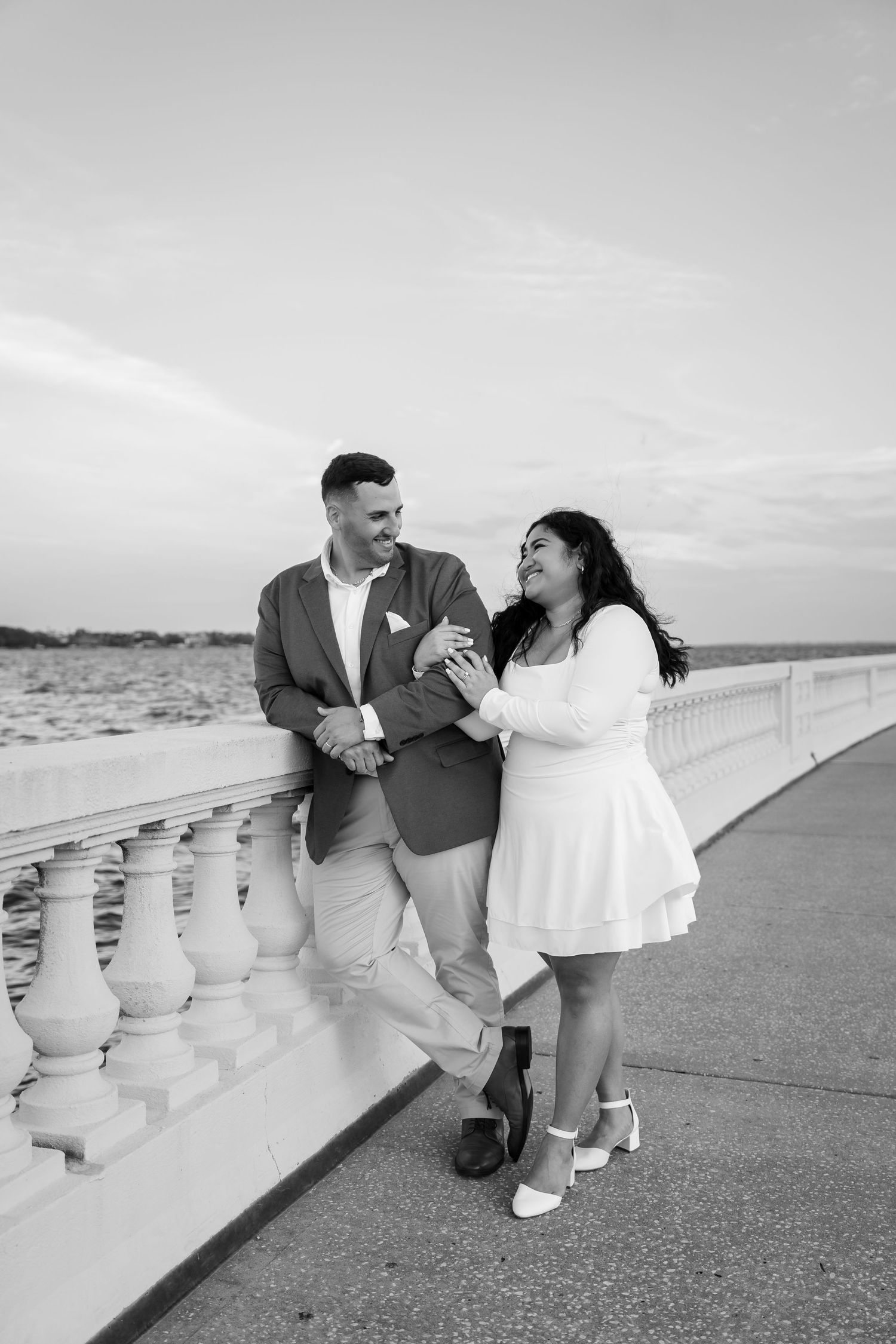 Black and white photo of a couple holding hands and smiling on a waterfront promenade at sunset.